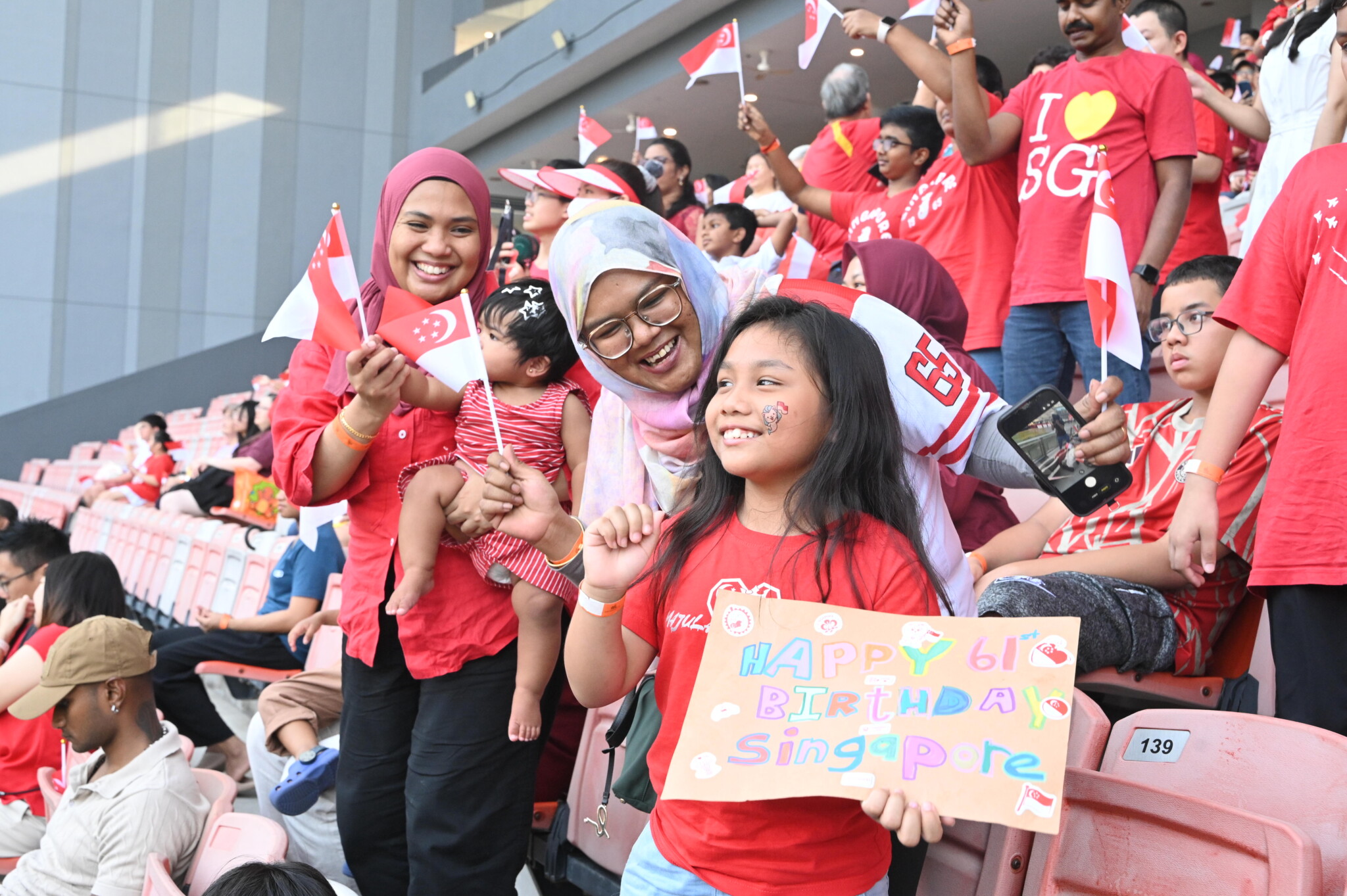 Crowd in red and white with a girl holding a handmade sign reading Happy 61st Birthday Singapore