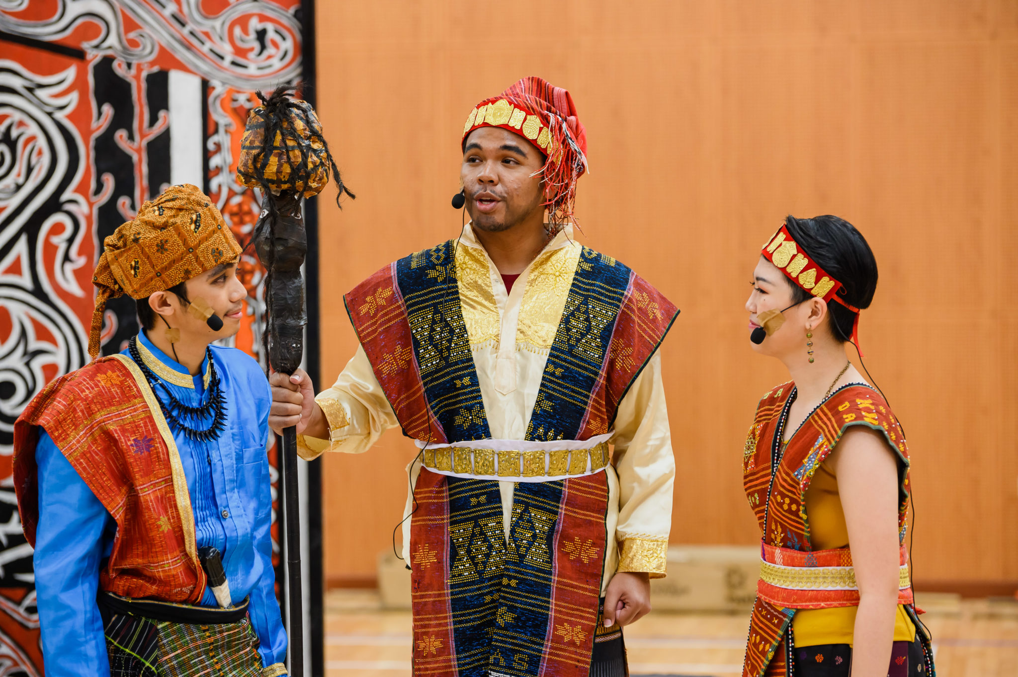 Three cast members in colourful Batak costumes in front of a carved backdrop