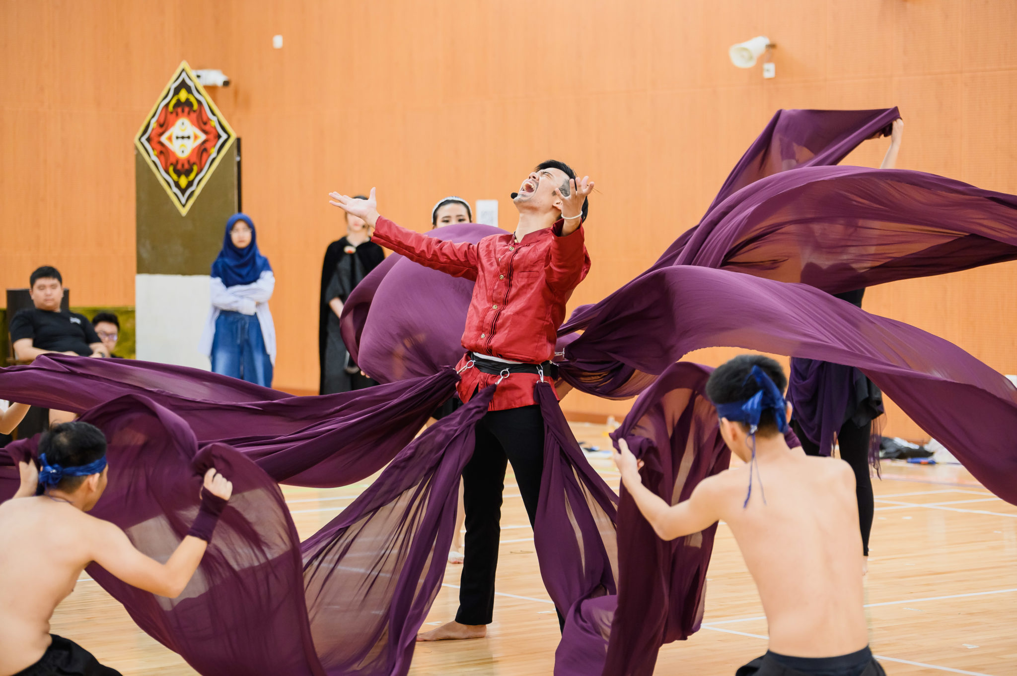 A performer in red holding up a large purple cloth while dancers move below at a rehearsal