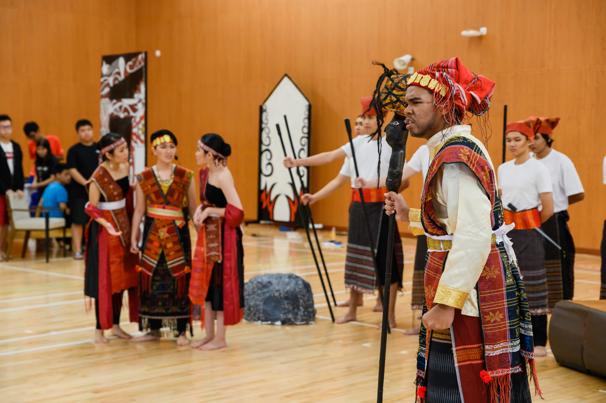 Cast members in Batak costumes holding staffs and waiting on the rehearsal floor