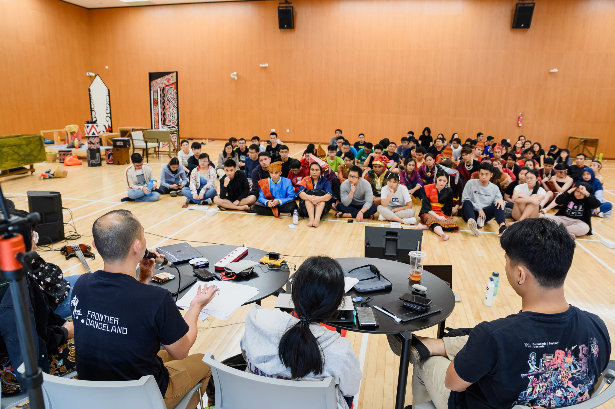 Cast and crew seated on the rehearsal hall floor while production staff work at a laptop table