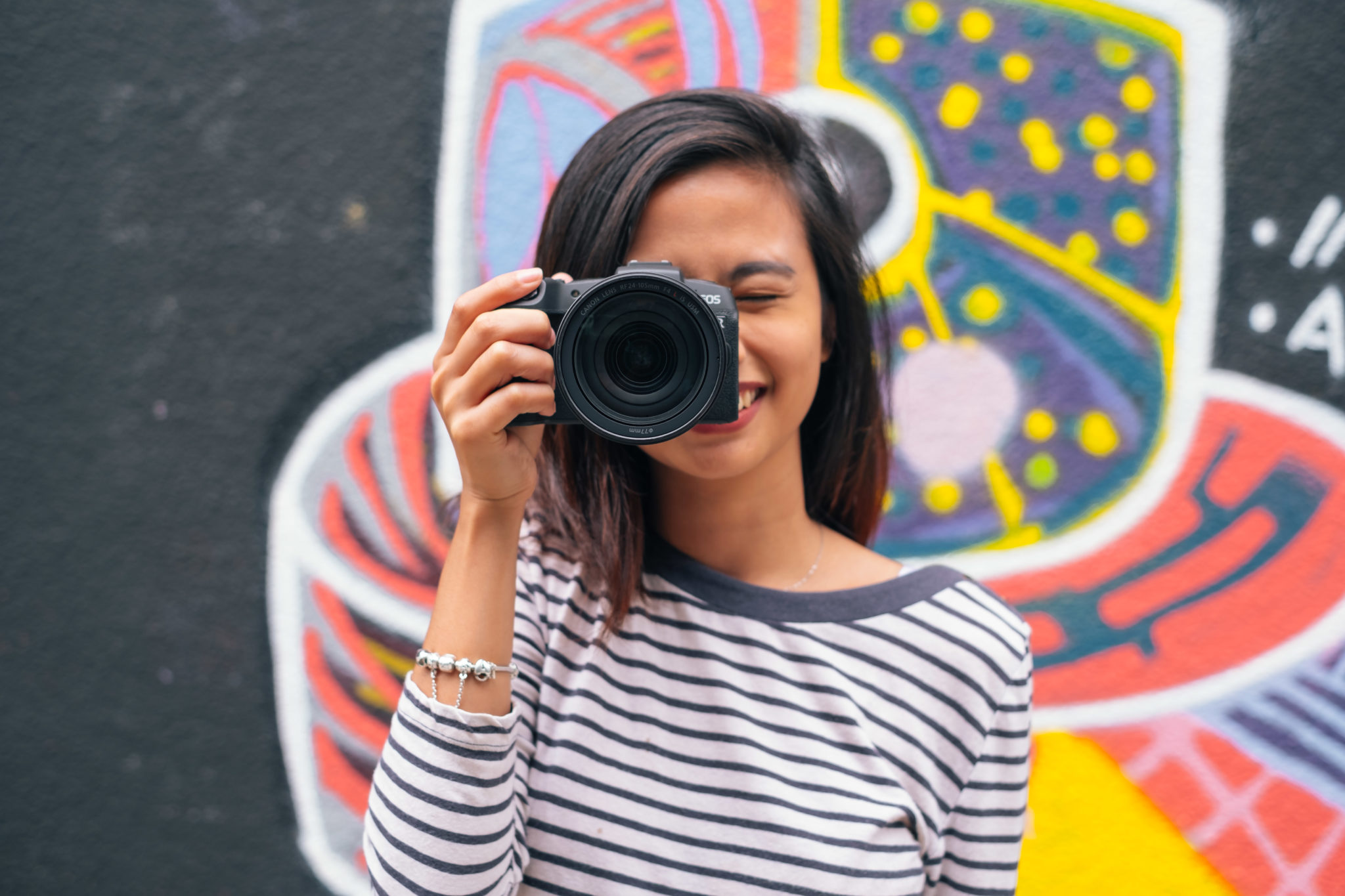 A woman in a striped top holding a camera up to her eye in front of a colourful mural