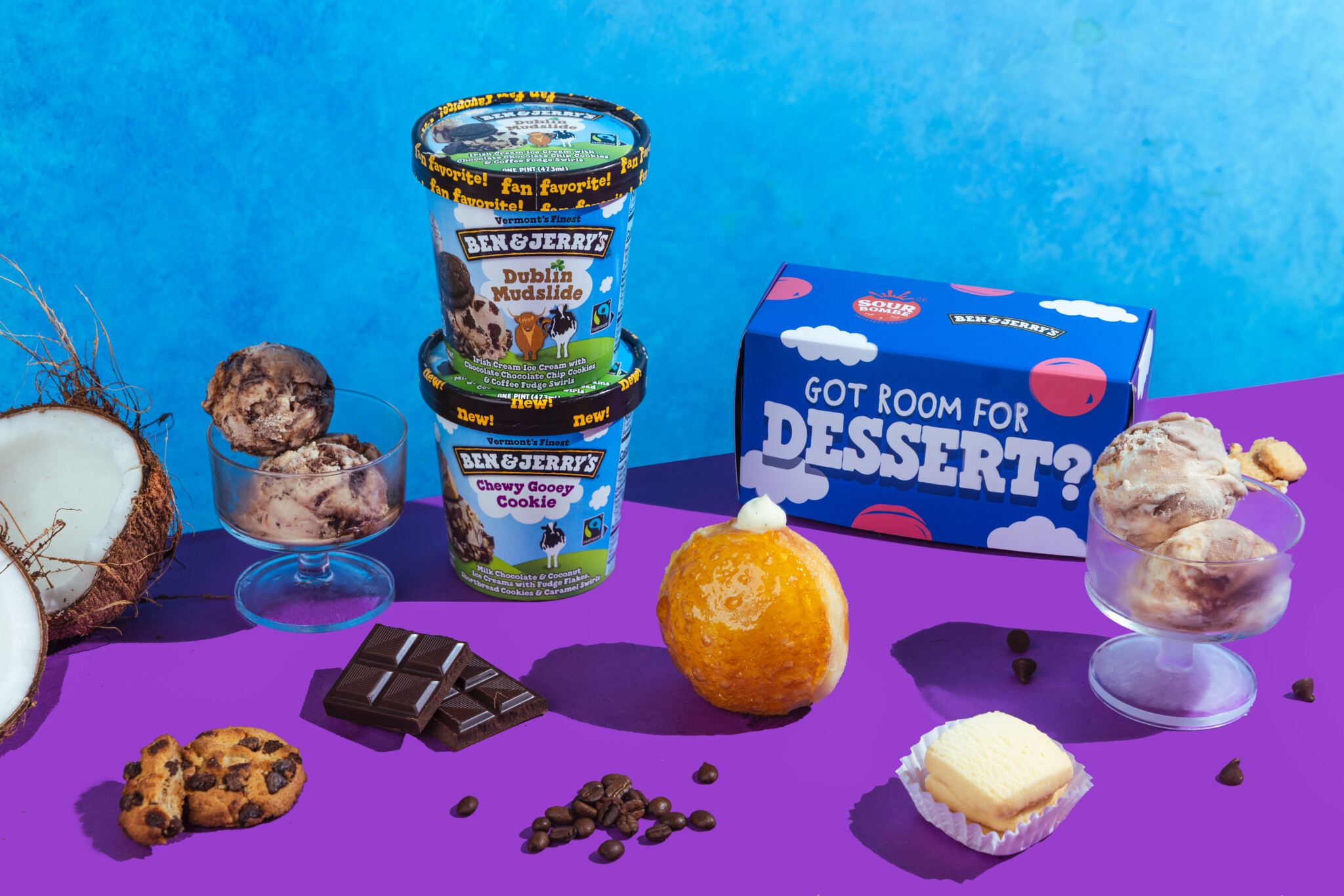 Two pints of Ben & Jerry's with a Brûlée Hooray doughnut and dessert props on a purple and blue backdrop