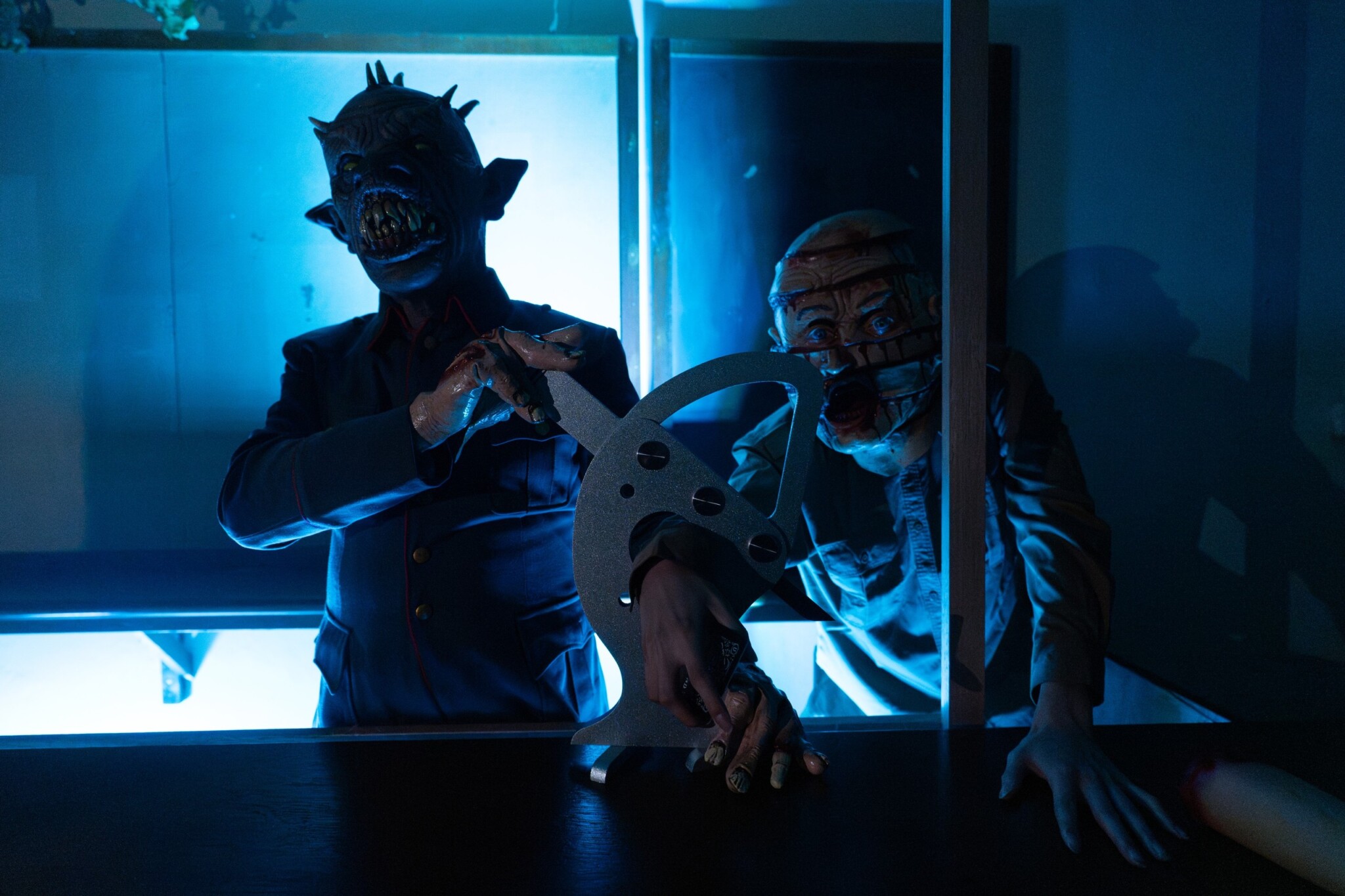 Two masked horror performers stand in a blue-lit room, one holding a saw prop.