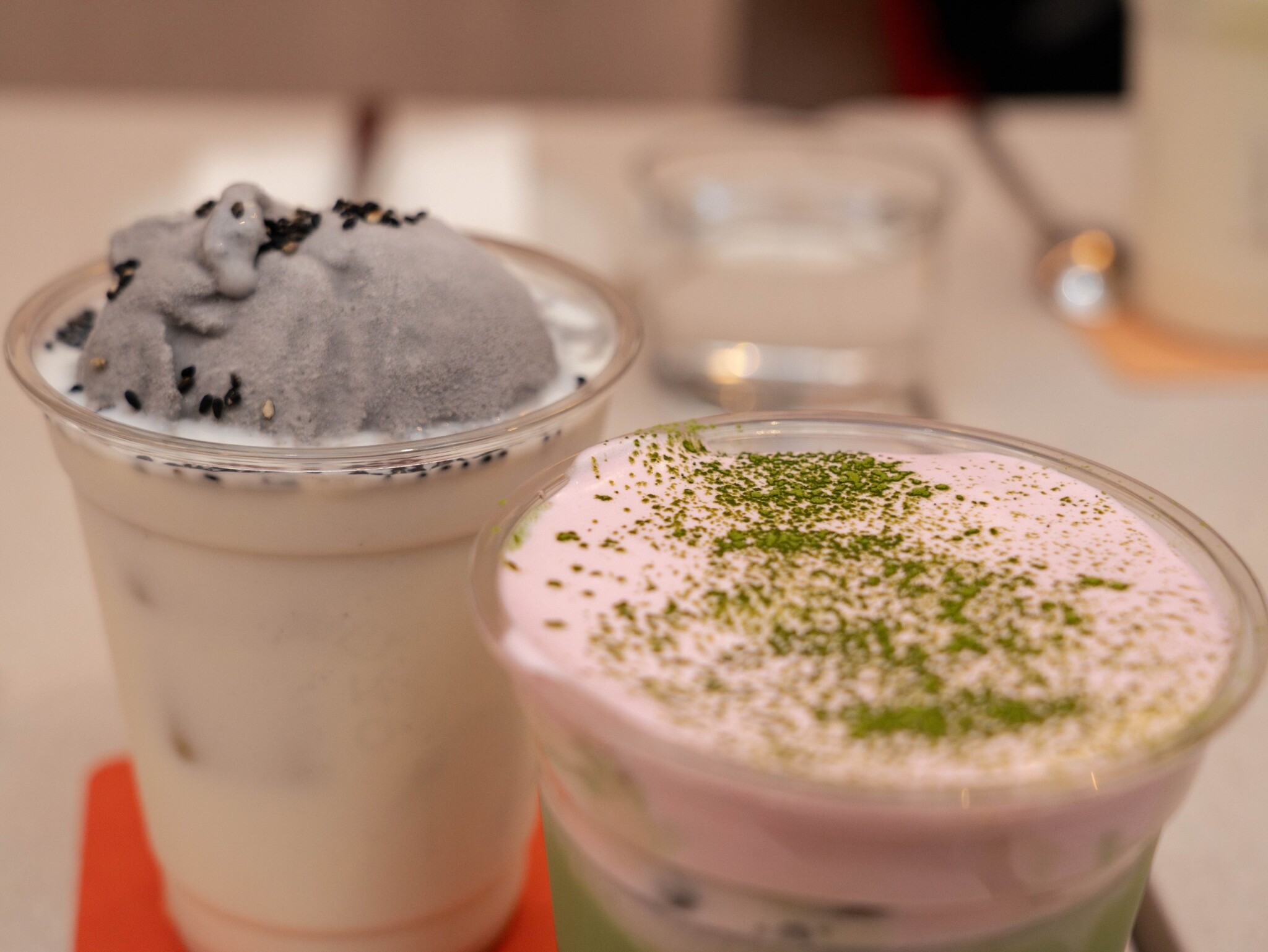 Two iced drinks sit on a table, one with dark sesame topping and one pink and green.