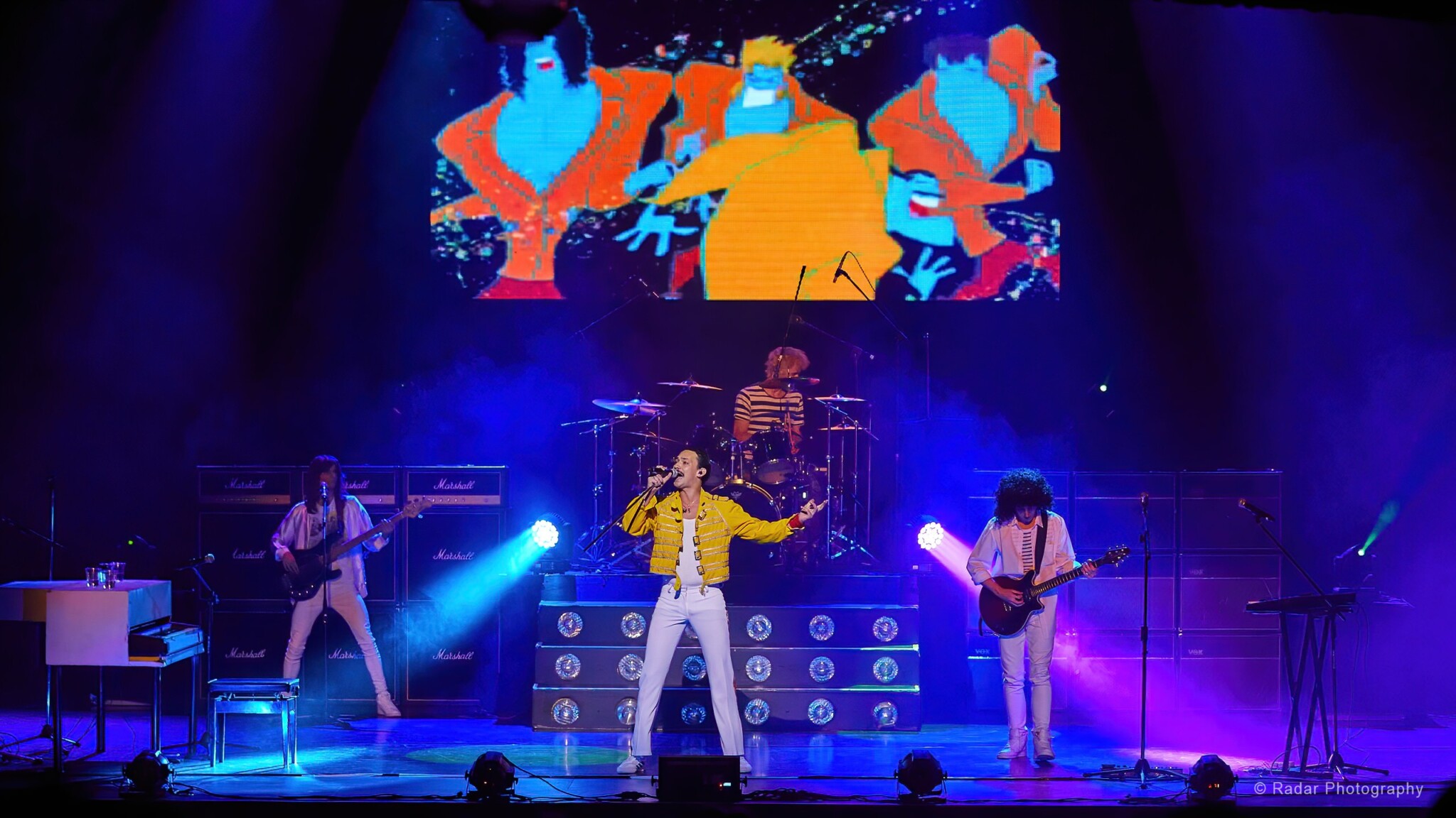 A singer in a yellow jacket performs on stage with the Queen tribute band.