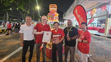 Five people pose outdoors at night next to a tiered 'Happy Birthday Singapore' cake display, with two of them holding a