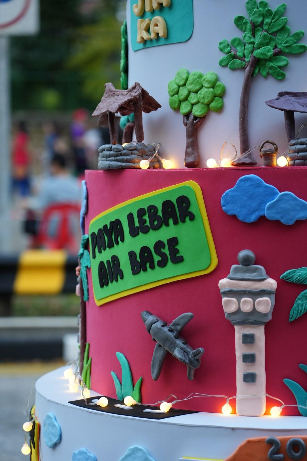 A cake tier with fondant details including a green Paya Lebar Air Base sign, a control tower, an airplane and string lights