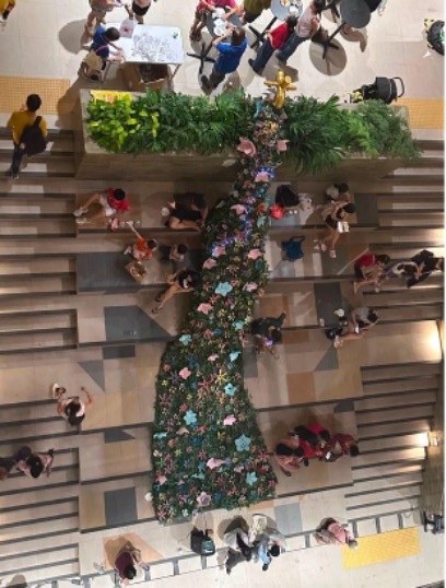 An aerial view of a large decorated tree with colourful ornaments in a mall atrium, with people on the surrounding staircases