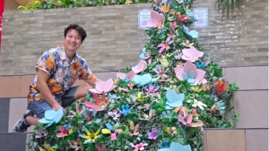 A man in a floral shirt kneels beside a wall installation covered in greenery and colourful paper flowers