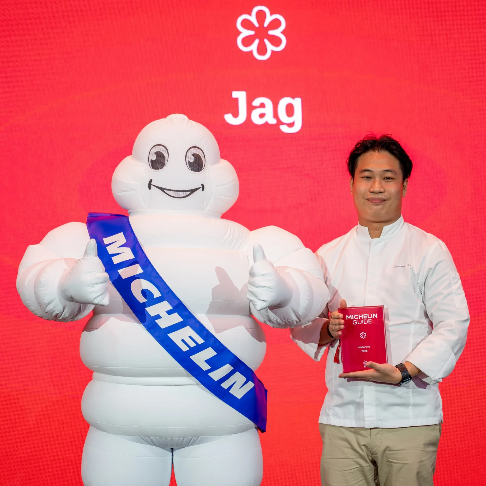 A chef in whites posing with the Michelin Man and the red MICHELIN Guide Singapore 2026 book on a JAG-branded backdrop