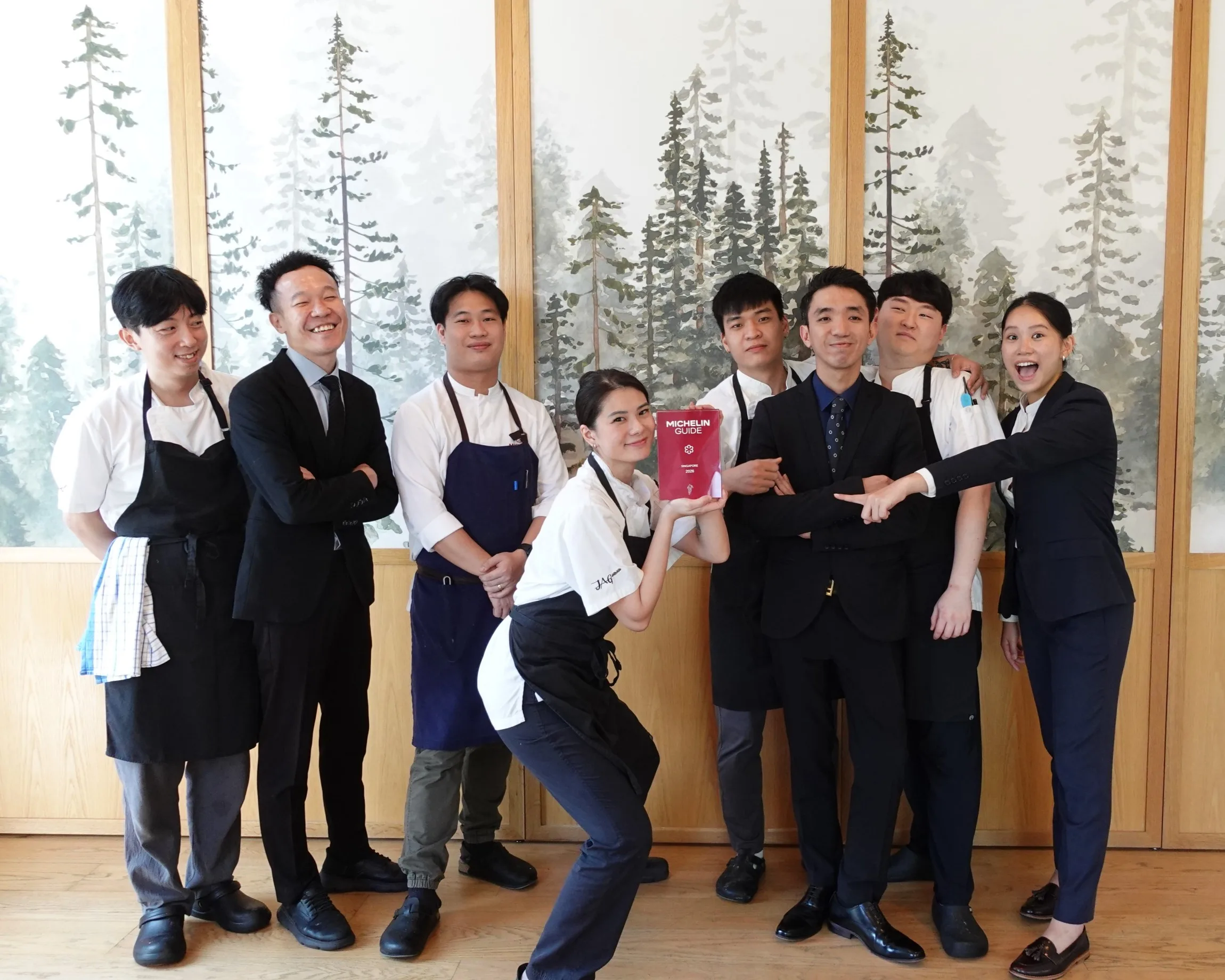 Eight JAG staff in aprons and suits posing together with the red 2026 guide in front of a forest mural