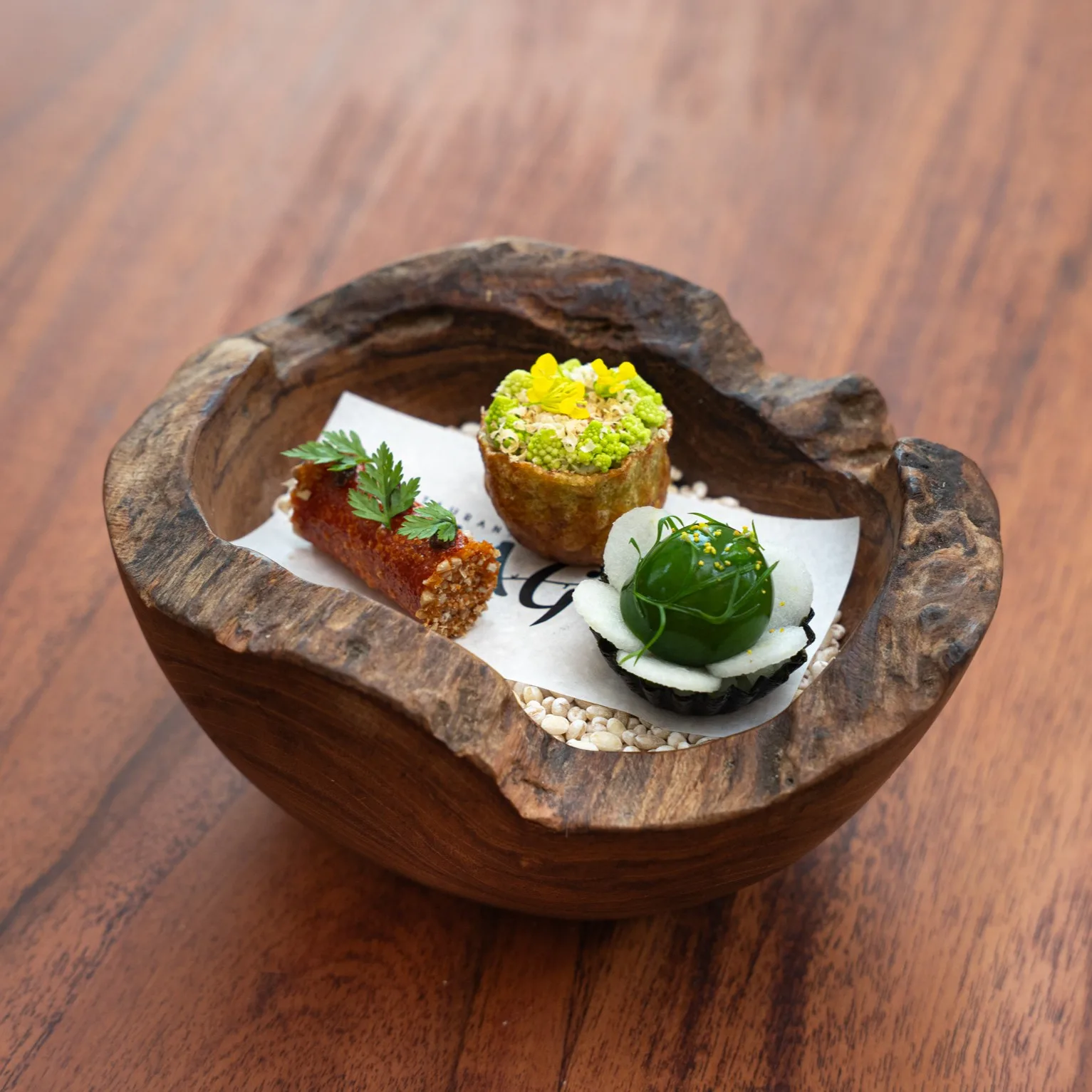 Three fine-dining canapés in a rustic wooden bowl on a branded paper liner over rice grains