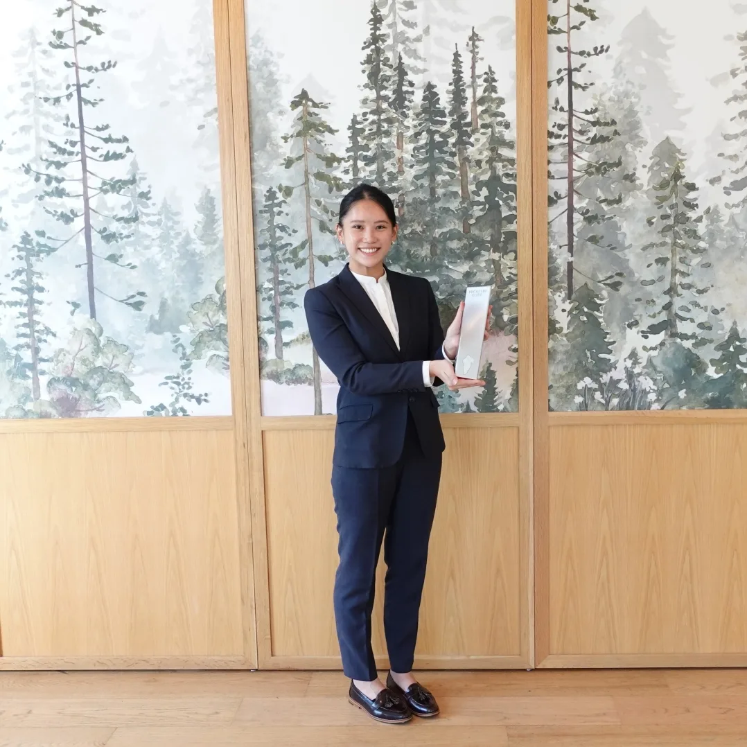 A woman in a navy suit smiling in front of a forest-themed mural, holding a silver box-shaped item