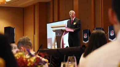 A man in a dark suit and red tie speaks at a podium bearing the Marina Bay Sands Singapore logo, addressing a seated audience