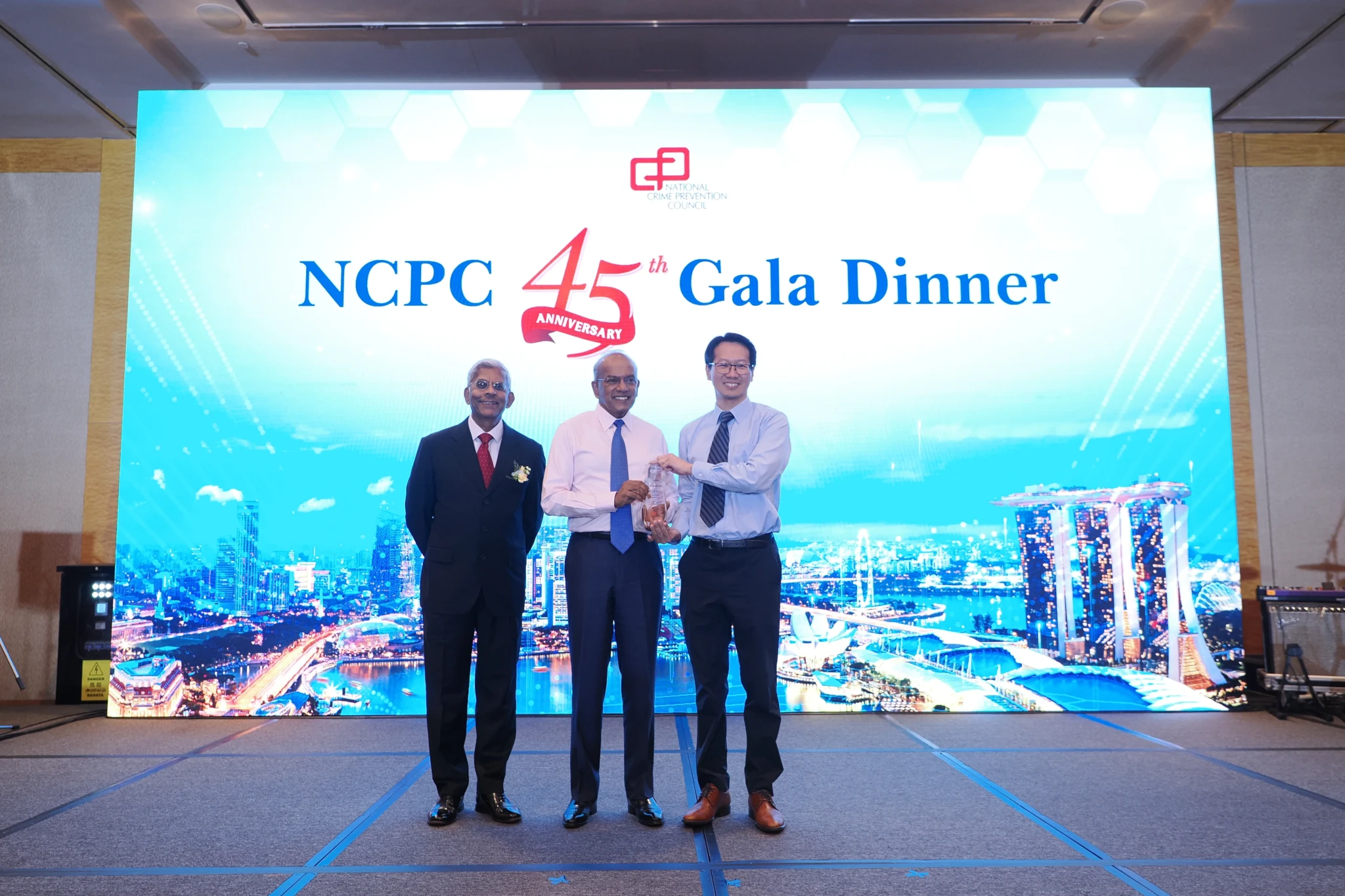 Three men in business attire stand on stage holding a trophy together at the NCPC 45th Anniversary Gala Dinner