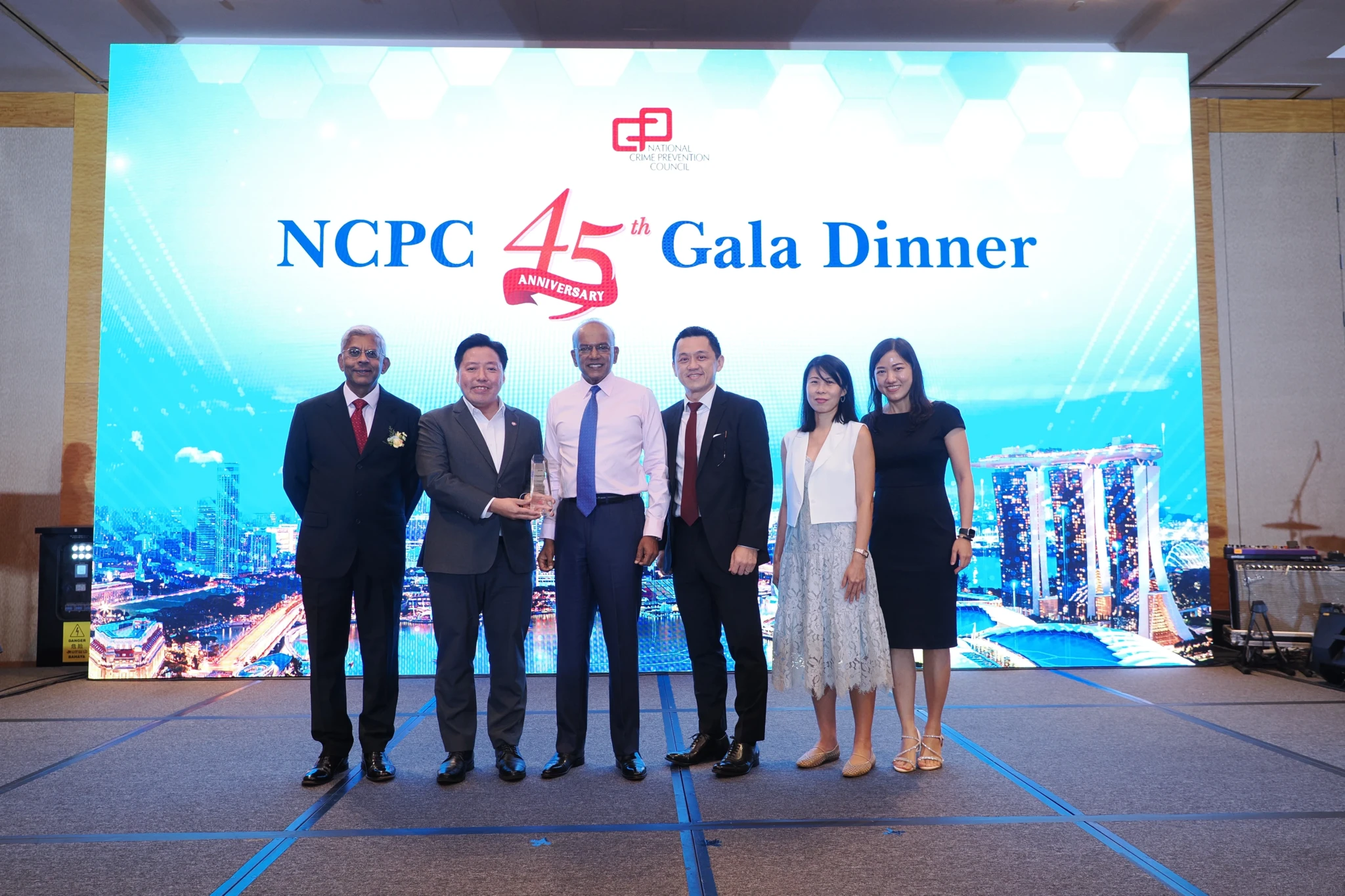 Six people in business attire stand on stage holding an award trophy at the NCPC 45th Anniversary Gala Dinner