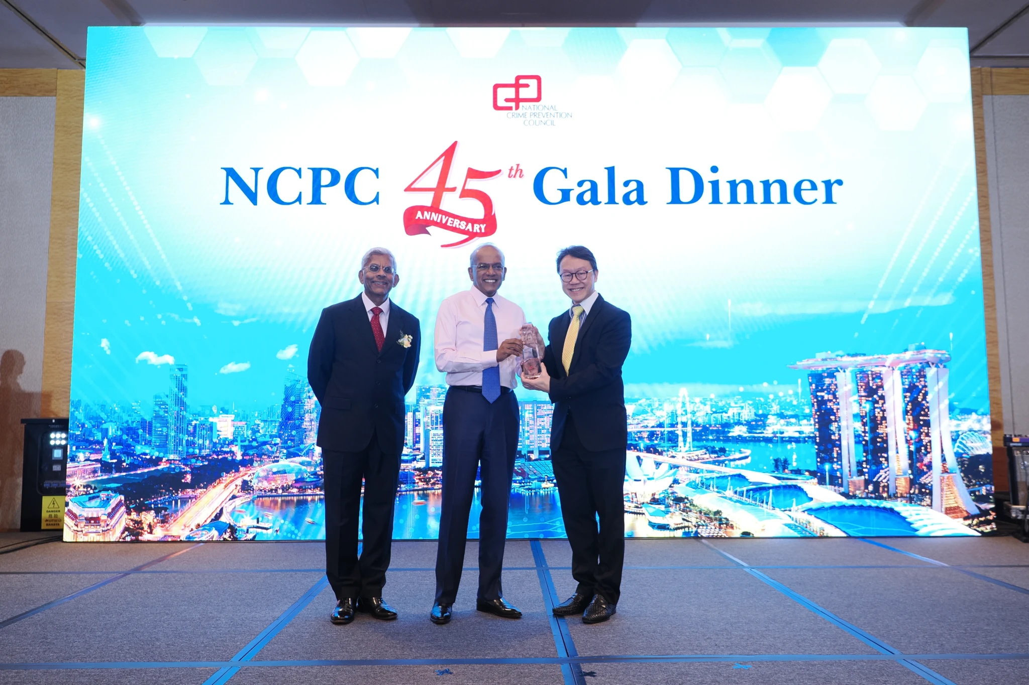 Three men in suits on stage, two of them jointly holding a glass award, at the NCPC 45th Anniversary Gala Dinner