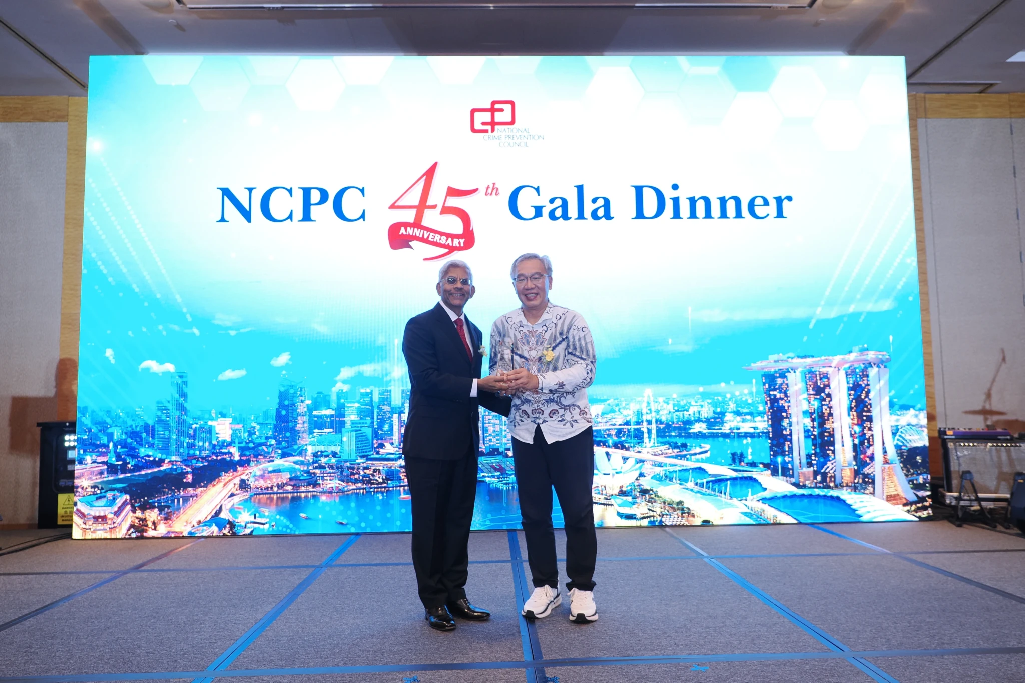 Two men in formal attire shake hands and hold an award on stage at the NCPC 45th Anniversary Gala Dinner