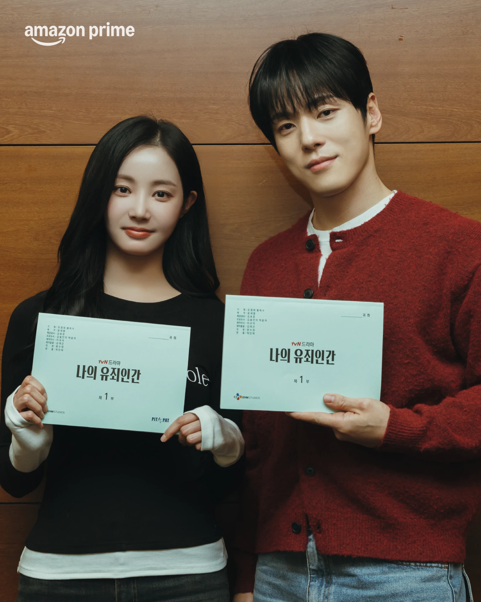 A woman in a black top and a man in a red cardigan hold Korean scripts against a wood-panelled wall