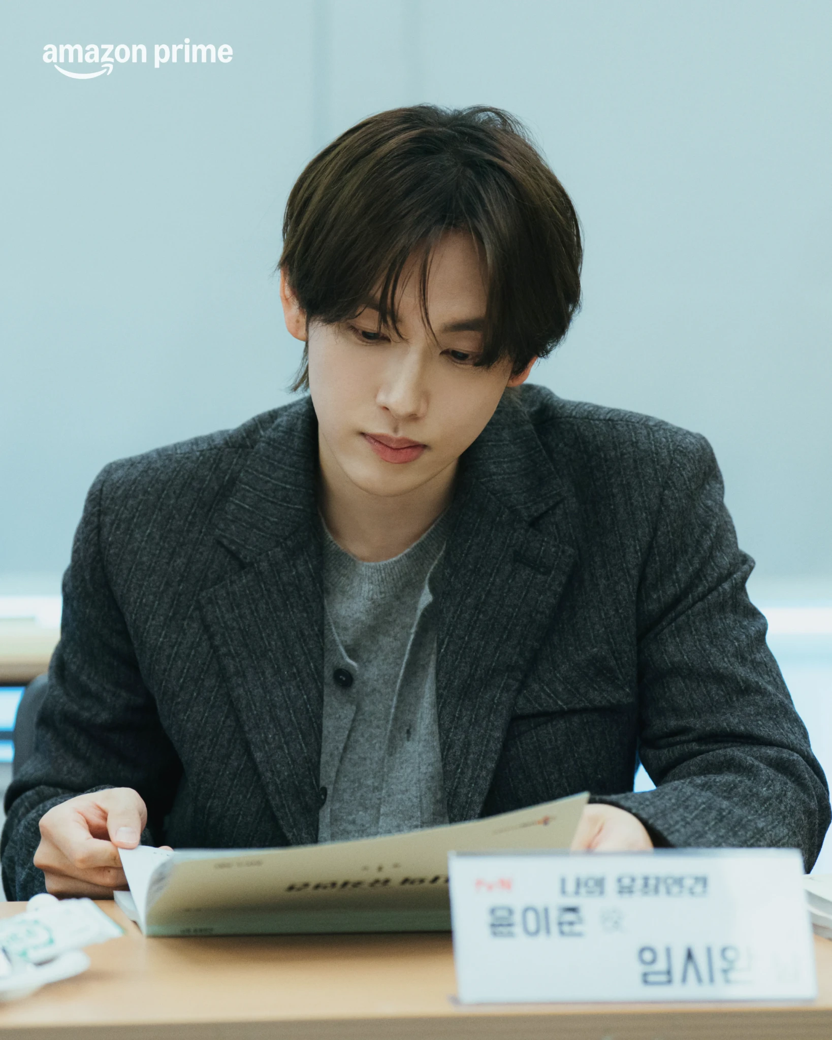 A man in a grey tweed blazer sits at a desk reading a script beside a Korean nameplate