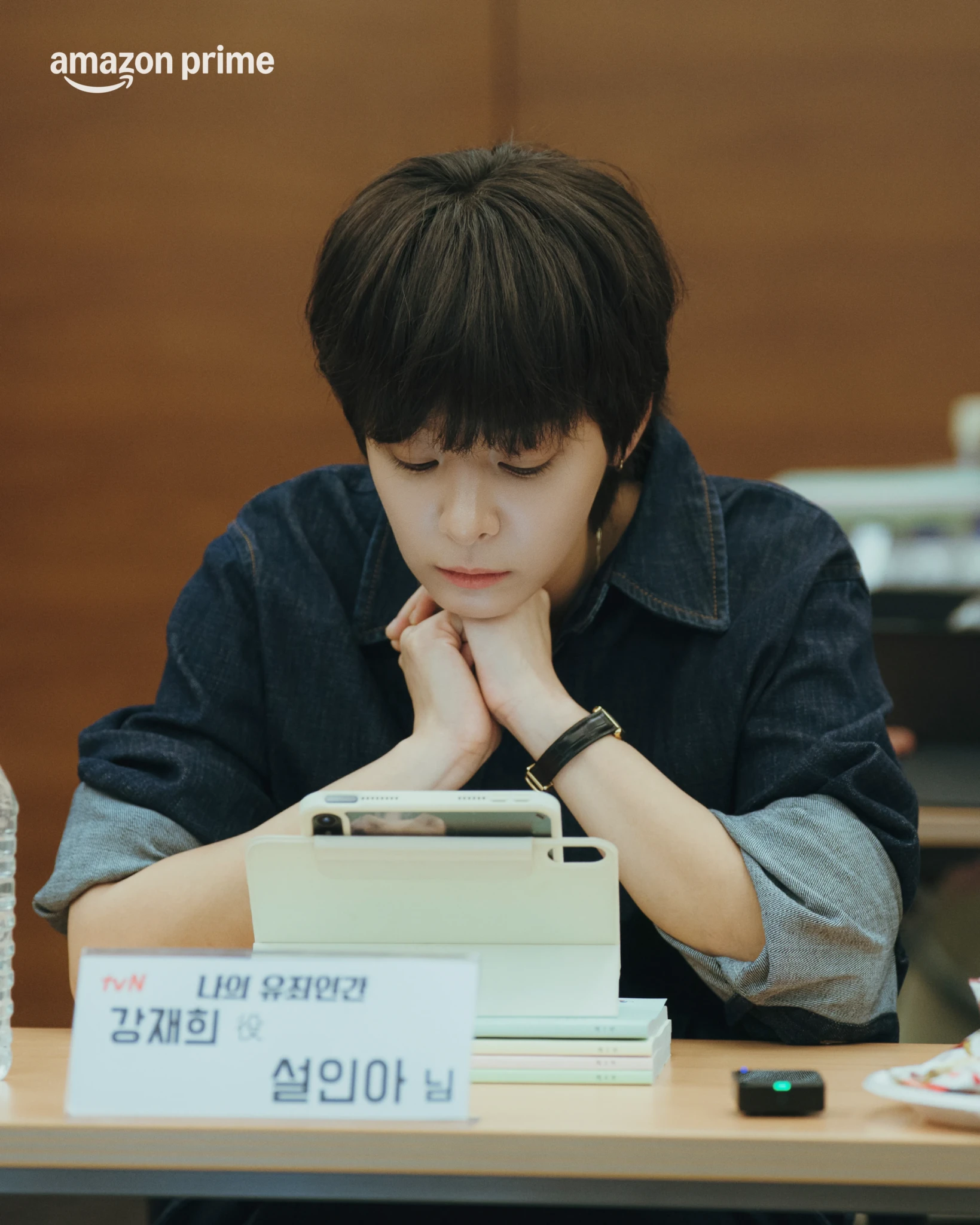 A young man with dark fringe rests his chin on his hands beside a name placard at the script reading