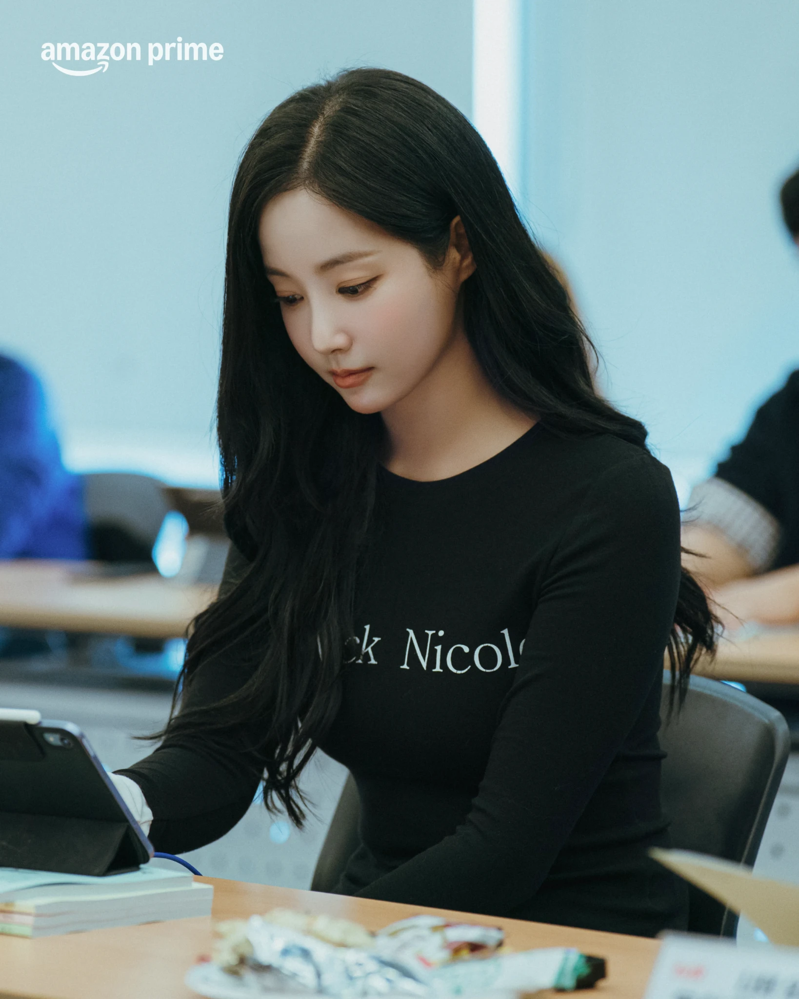 A young woman with long dark hair sits at a table using a tablet during the script reading