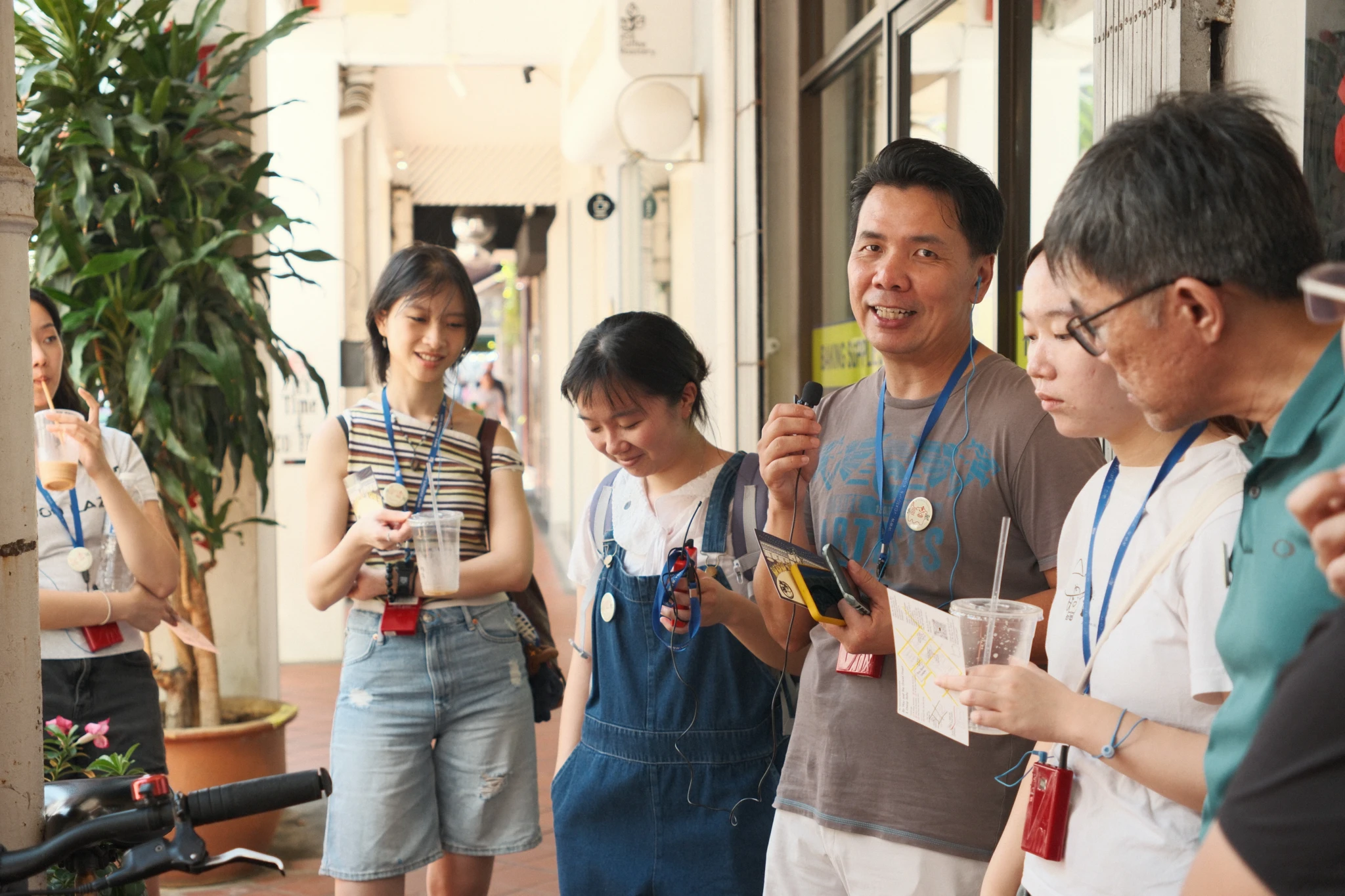 A guide speaks into a handheld microphone as a small group with lanyards and iced drinks listens under a shophouse five-foot way