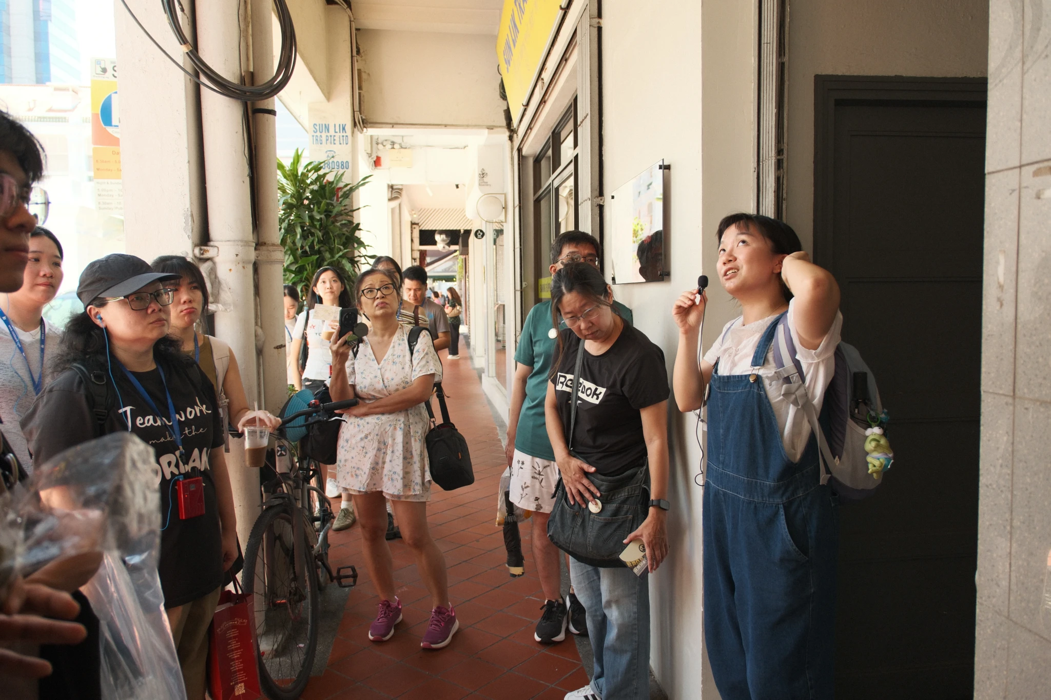 A guide in denim overalls addresses a walking tour group gathered in a shophouse five-foot way