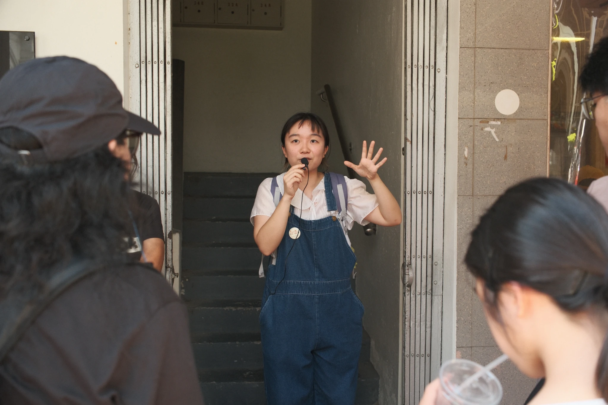 A tour guide in denim overalls speaks into a lapel microphone while addressing a group at a building's stairwell entrance