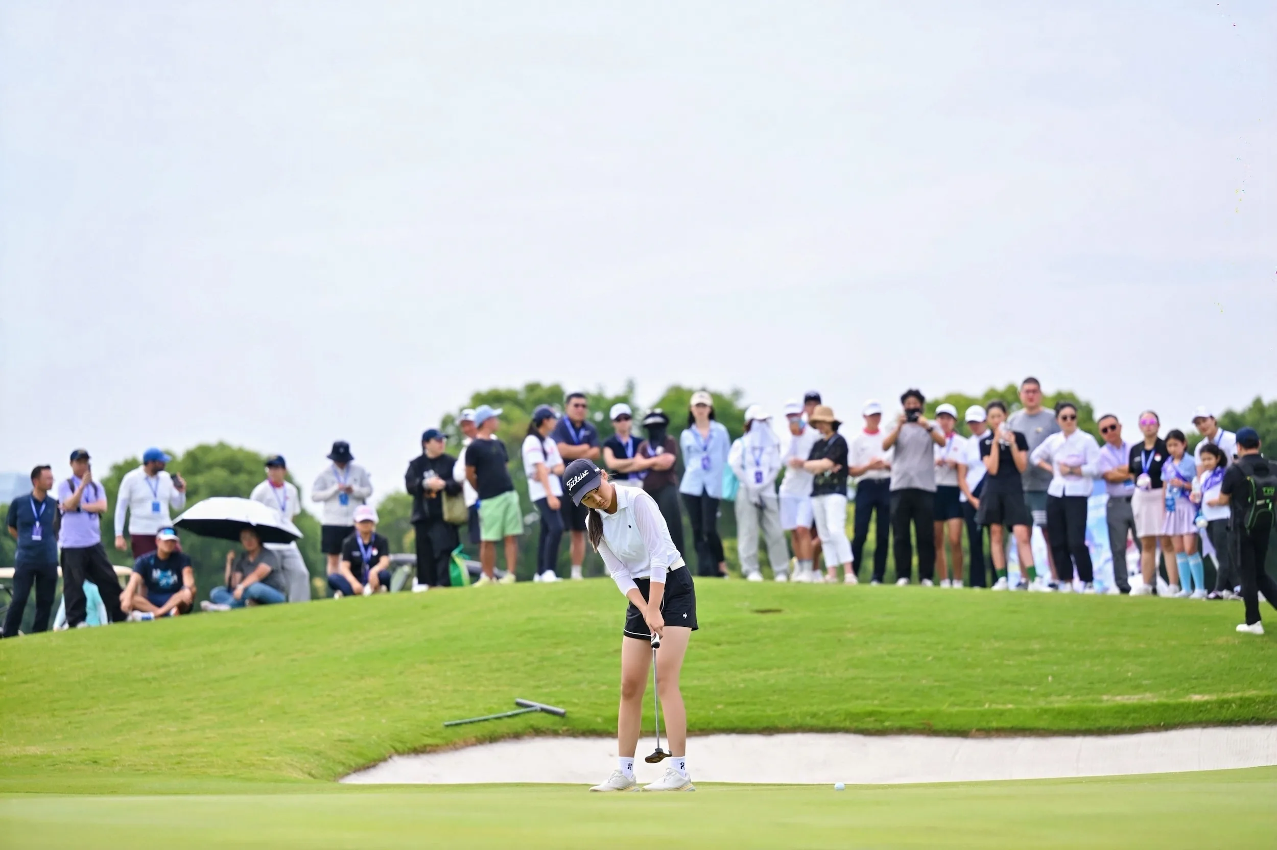 A junior golfer in a Titleist cap putts on the green as spectators watch