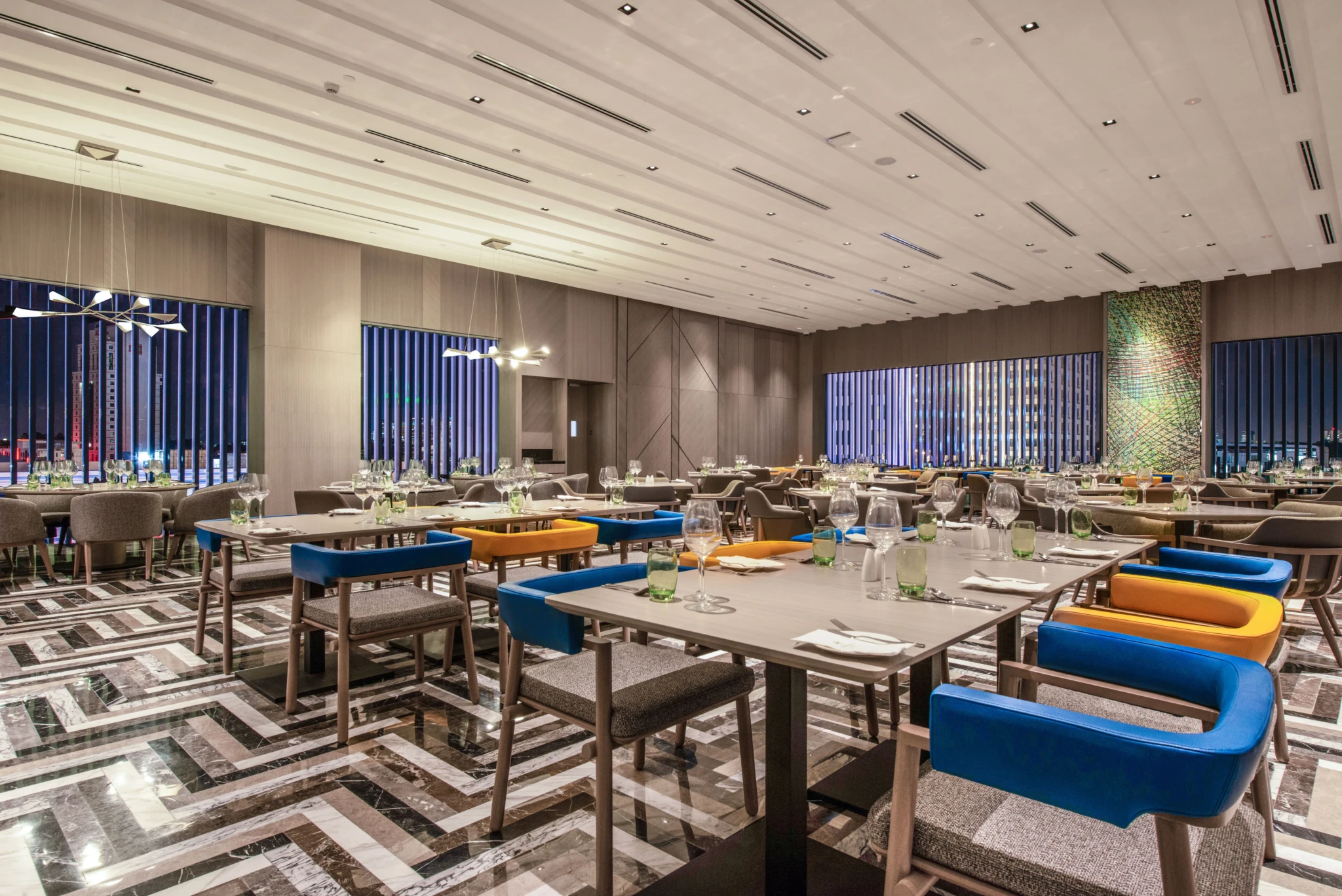Upscale restaurant dining room at night with marble herringbone flooring and a city skyline through slatted windows