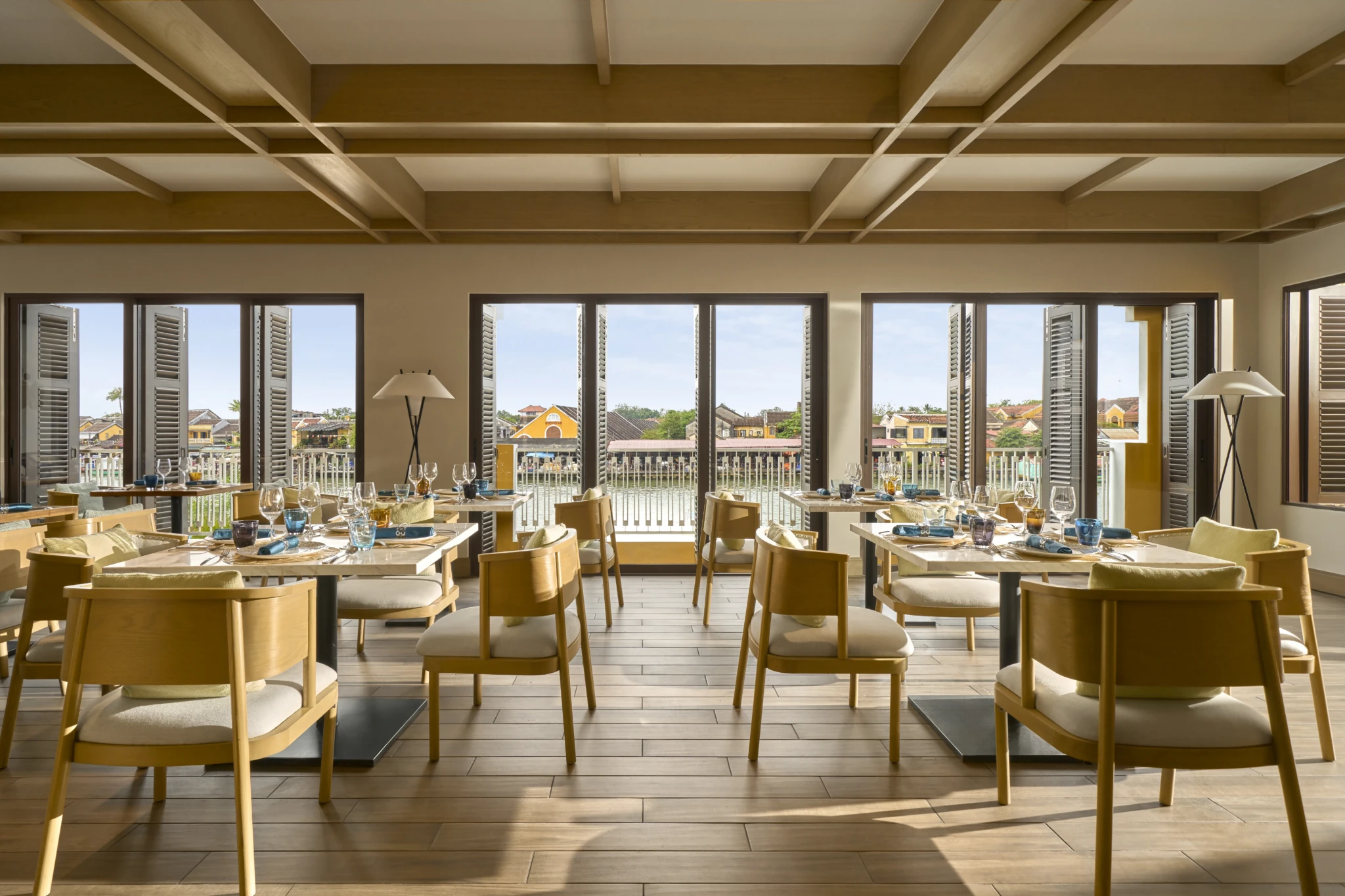 Sunlit hotel restaurant with wooden tables overlooking a riverside town through open shuttered doors