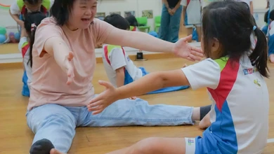 An Assistant Movement Co-facilitator guides a young preschooler through a stretching exercise on a classroom floor
