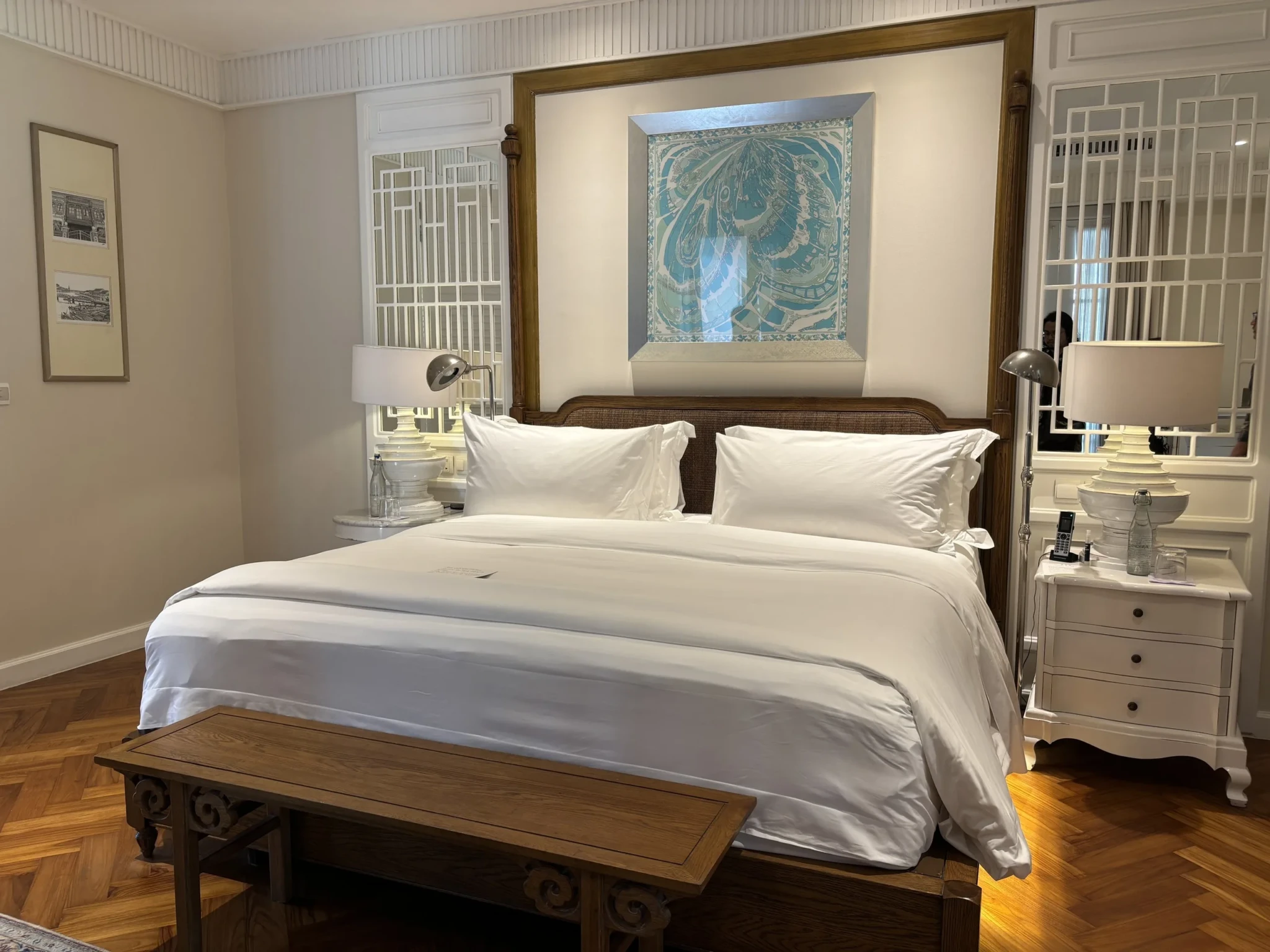 A bedroom with a wooden-framed bed, white linens, and blue framed artwork between white-shuttered windows