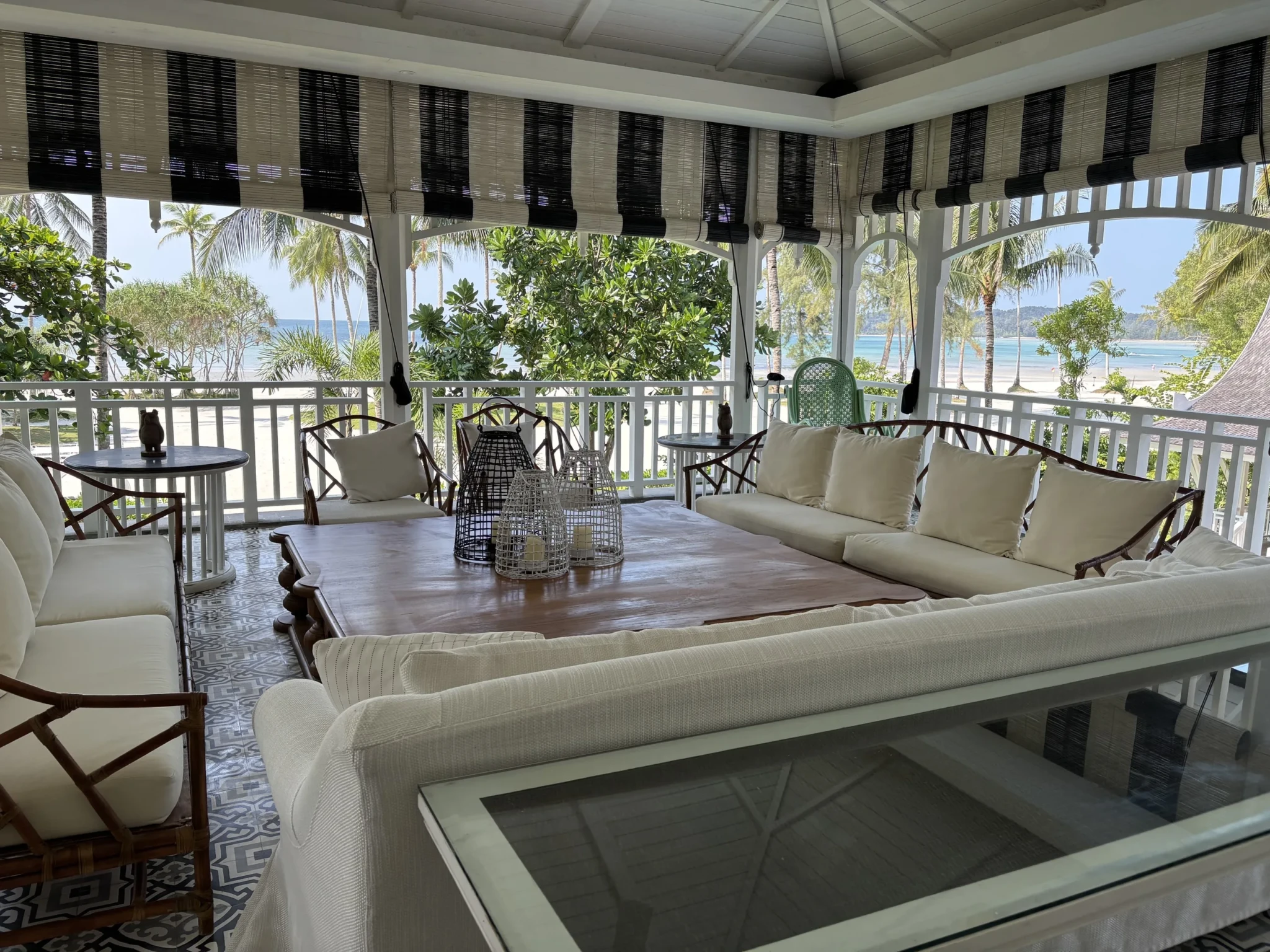 A covered beachfront pavilion with cream-upholstered seating and dark wooden chairs overlooking palms and the ocean
