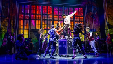 Performers from MJ The Michael Jackson Musical on stage under coloured lighting with an illuminated grid backdrop