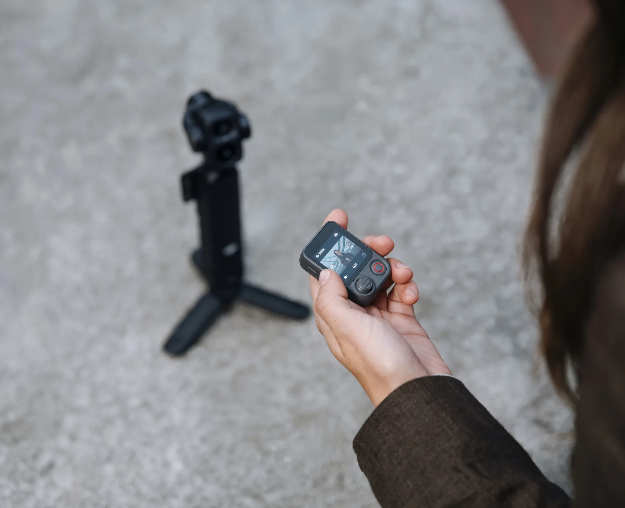 A hand holds the FrameTap remote with the DJI Osmo Pocket 4P on its tripod in the background