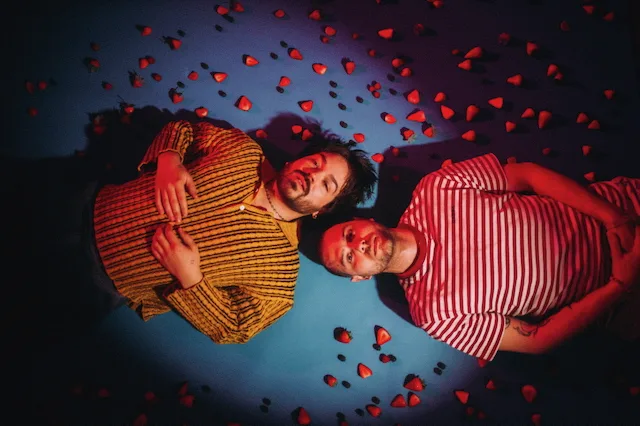 Milky Chance posing under falling red paper hearts against a blue background