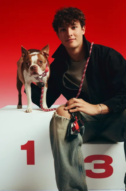 Ollie Bearman sits on a podium block beside his Boston Terrier Ruby against a red background