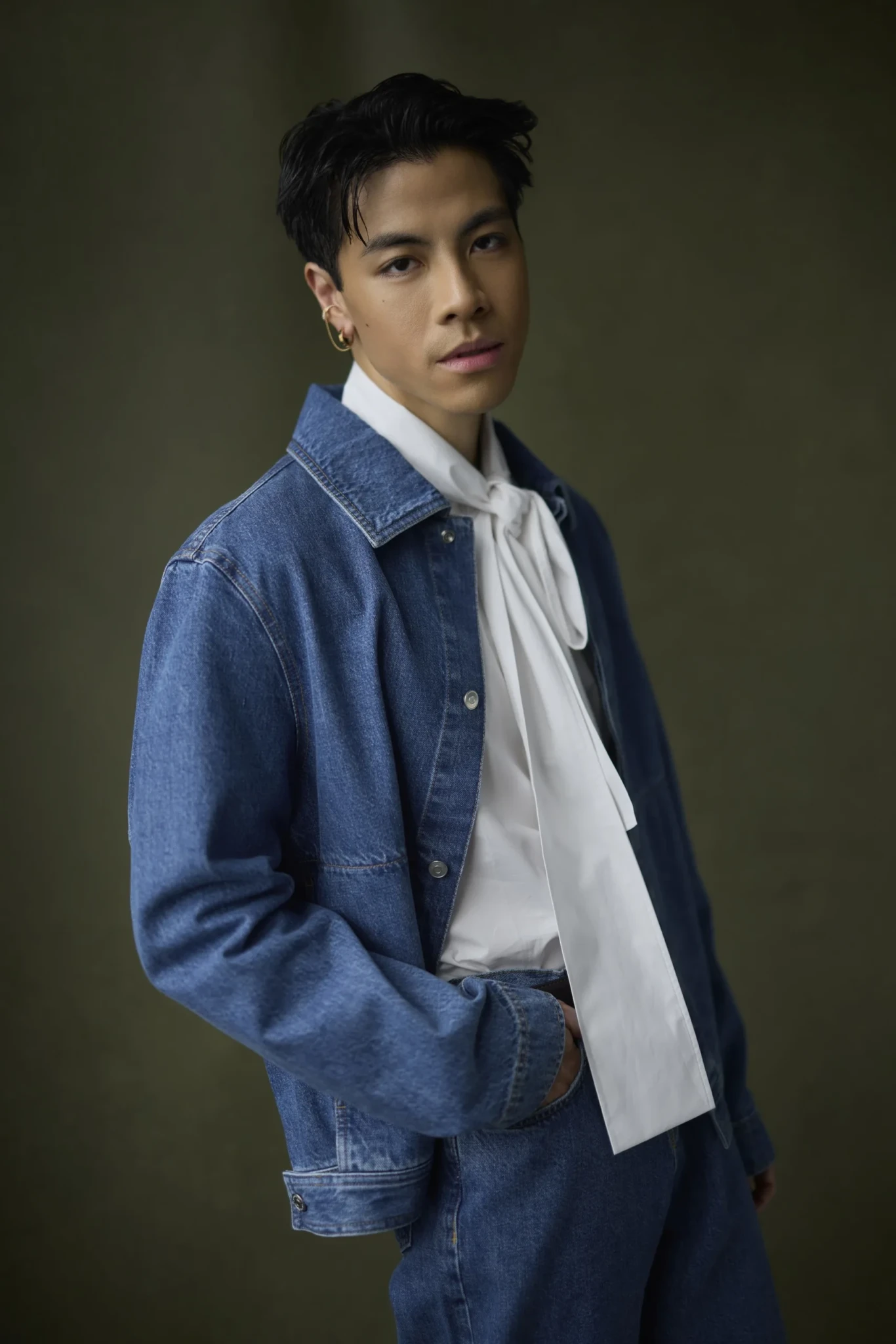 A young man in a white tie-neck blouse, open denim jacket, and denim trousers poses against a dark olive background