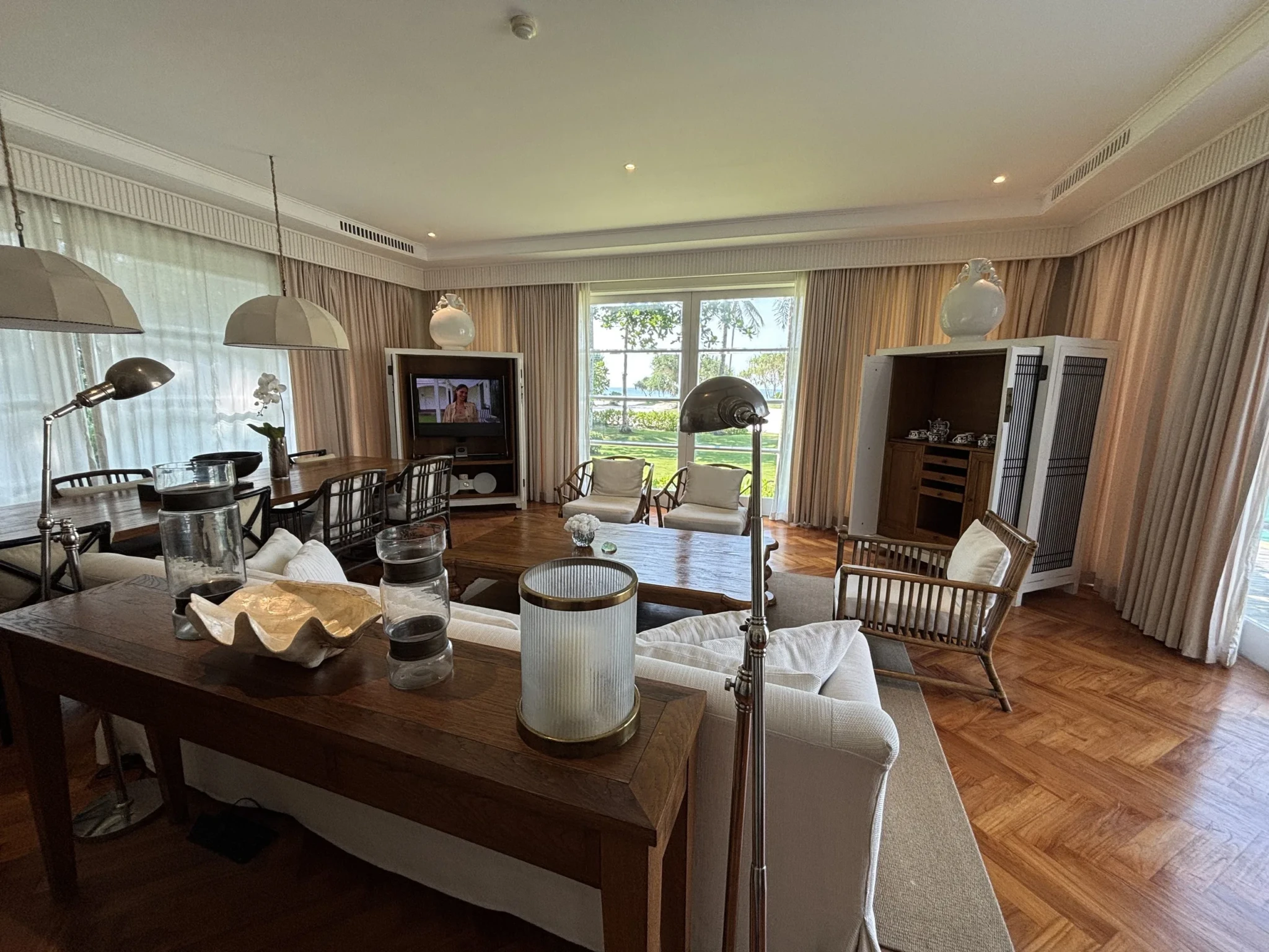 A suite living area with dark wood tables, cream seating, and ribbed-glass pendant lights by large windows