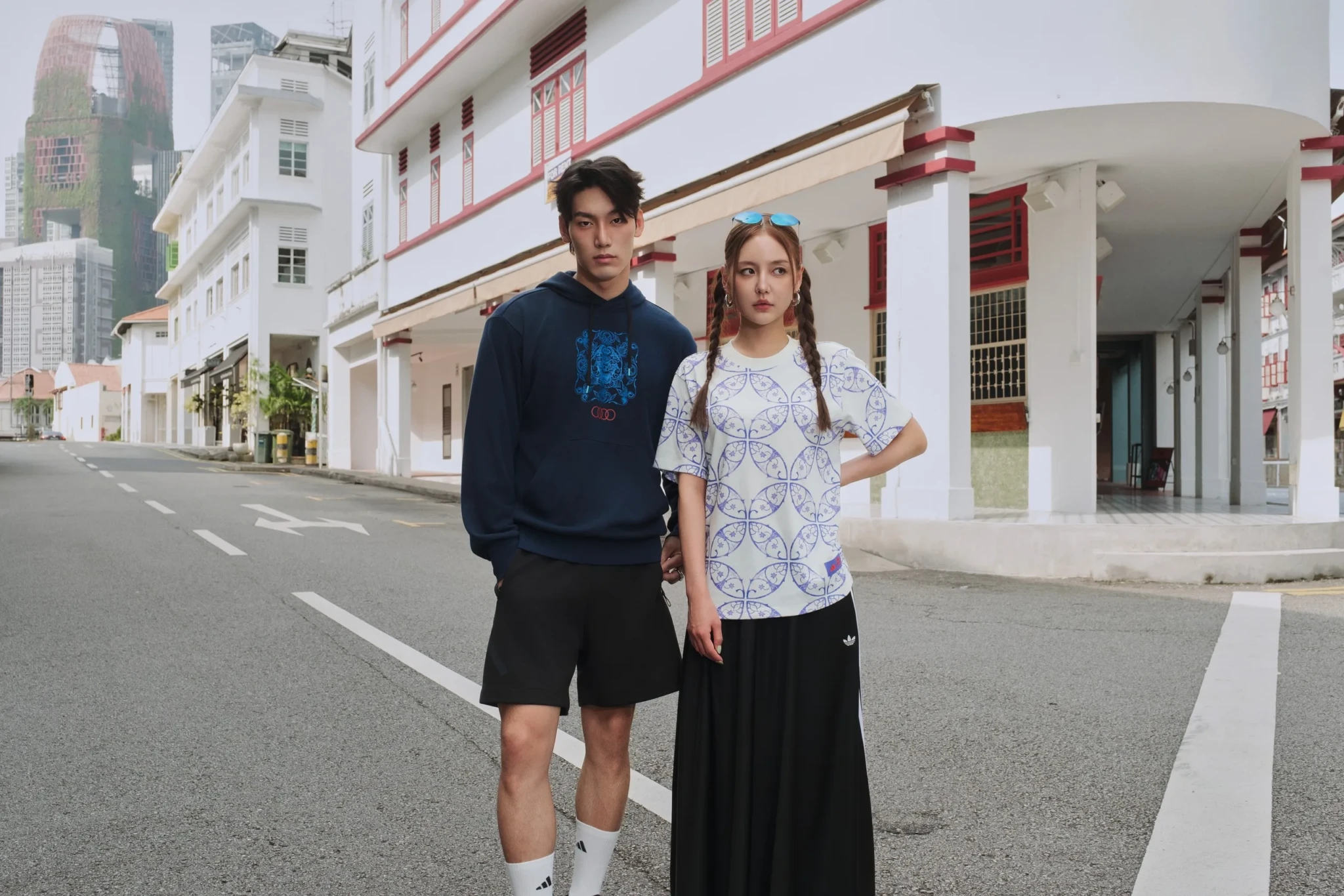 A man in a navy hoodie and a woman in a patterned T-shirt stand before white shophouses with red trim