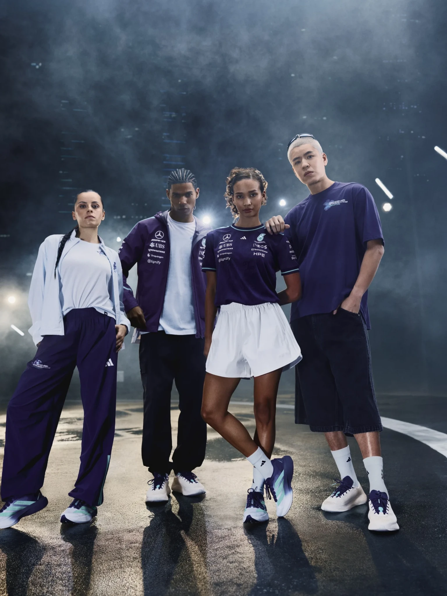 Four models stand on a wet track wearing purple and white adidas x Mercedes-AMG PETRONAS F1 Team apparel