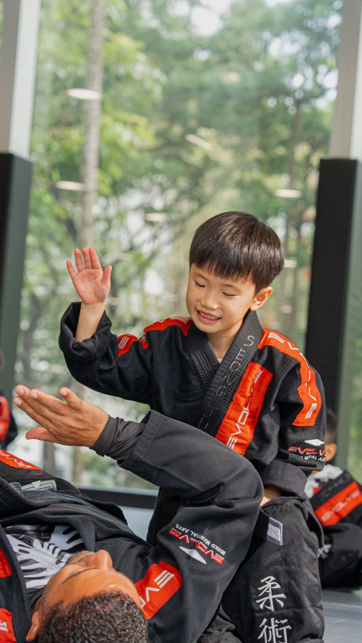 A child in a red-and-black martial arts uniform smiles and waves indoors