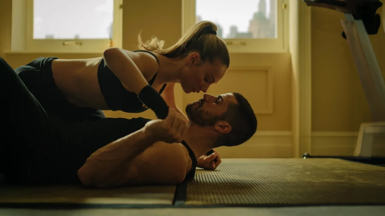 A man and a woman in workout attire face each other while doing an abdominal exercise on yoga mats in a sunlit room