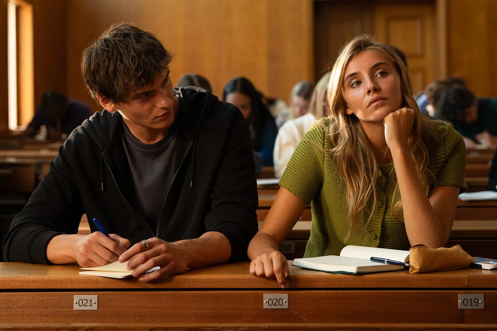 A young man in a hoodie and a young woman in a green cardigan share a desk in a lecture hall