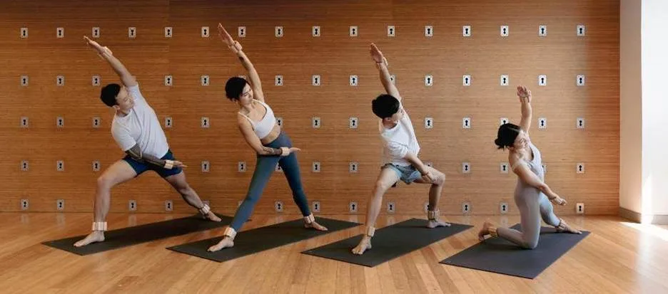 Four people hold an extended side-angle yoga pose on black mats in a wood-panelled studio