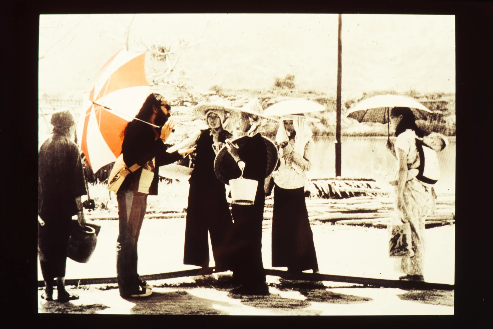 A man holding a red-and-white striped umbrella gestures towards a riverside crowd carrying baskets