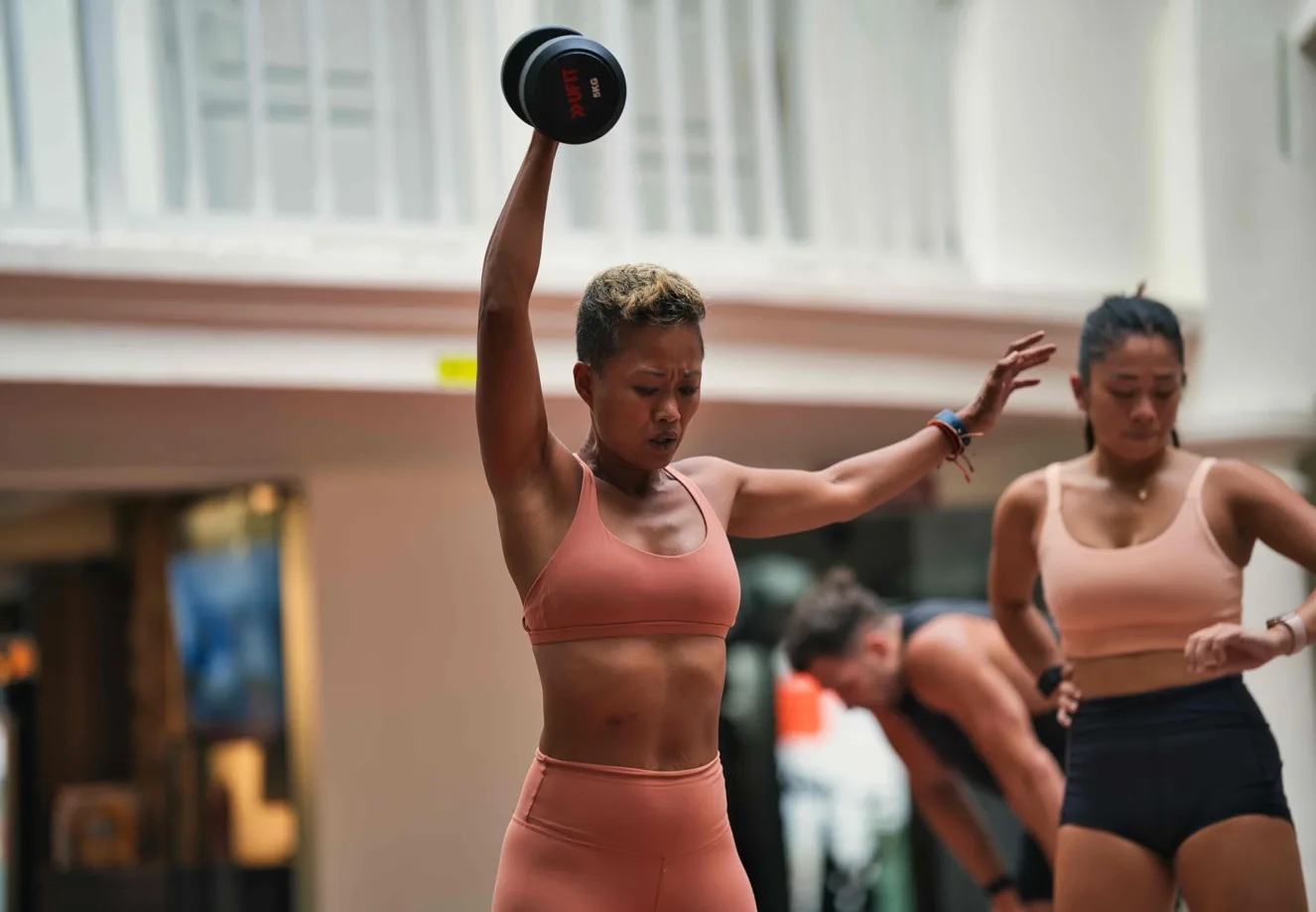 A woman in a peach sports bra and leggings raises a dumbbell overhead during an outdoor group fitness class, with two other participants visible behind her.